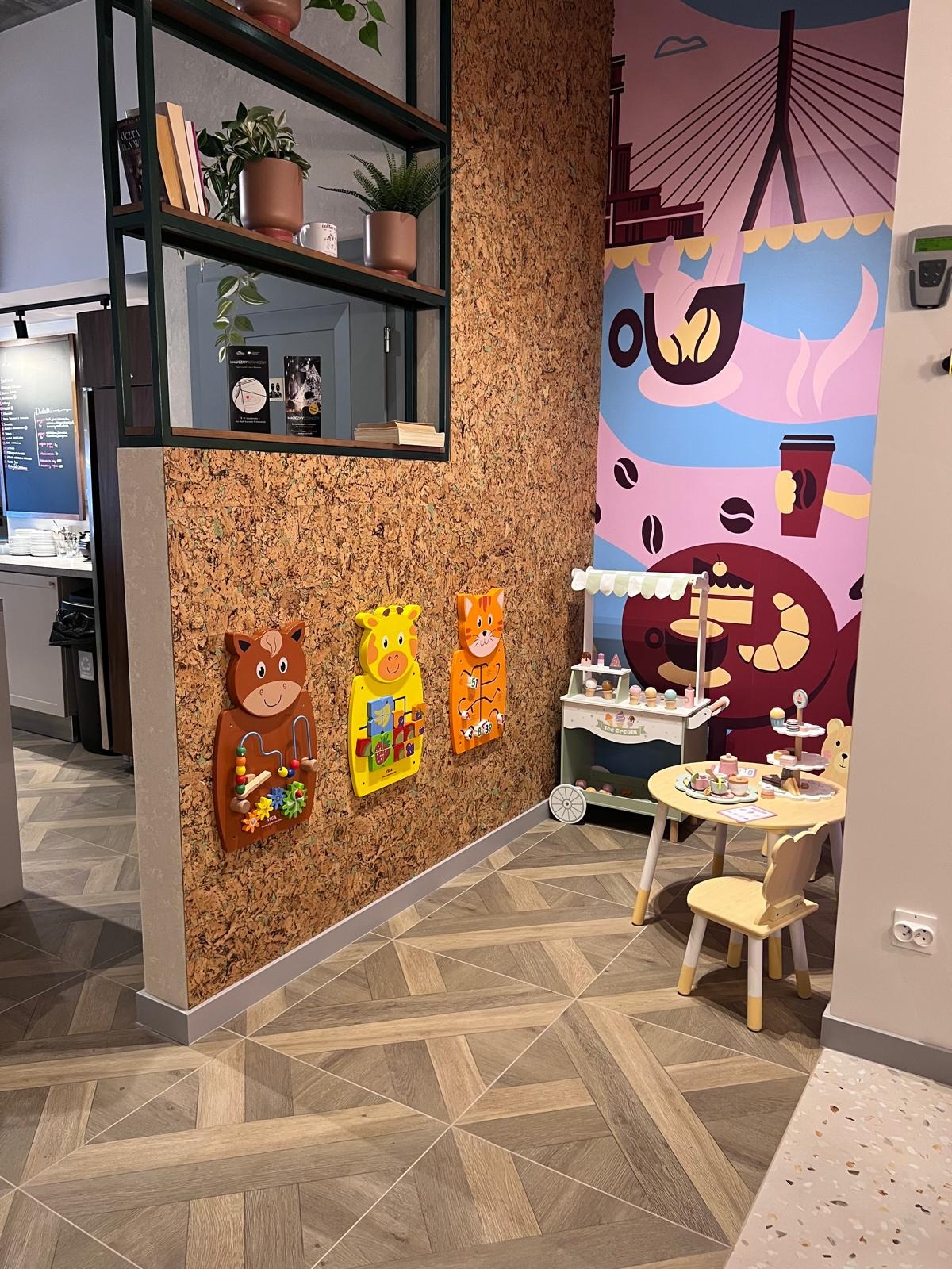 Kącik zabaw dla dzieci w kawiarni w Warszawie: drewniane zabawki na korkowej ścianie, stolik z krzesełkiem i mural z motywem kawy.