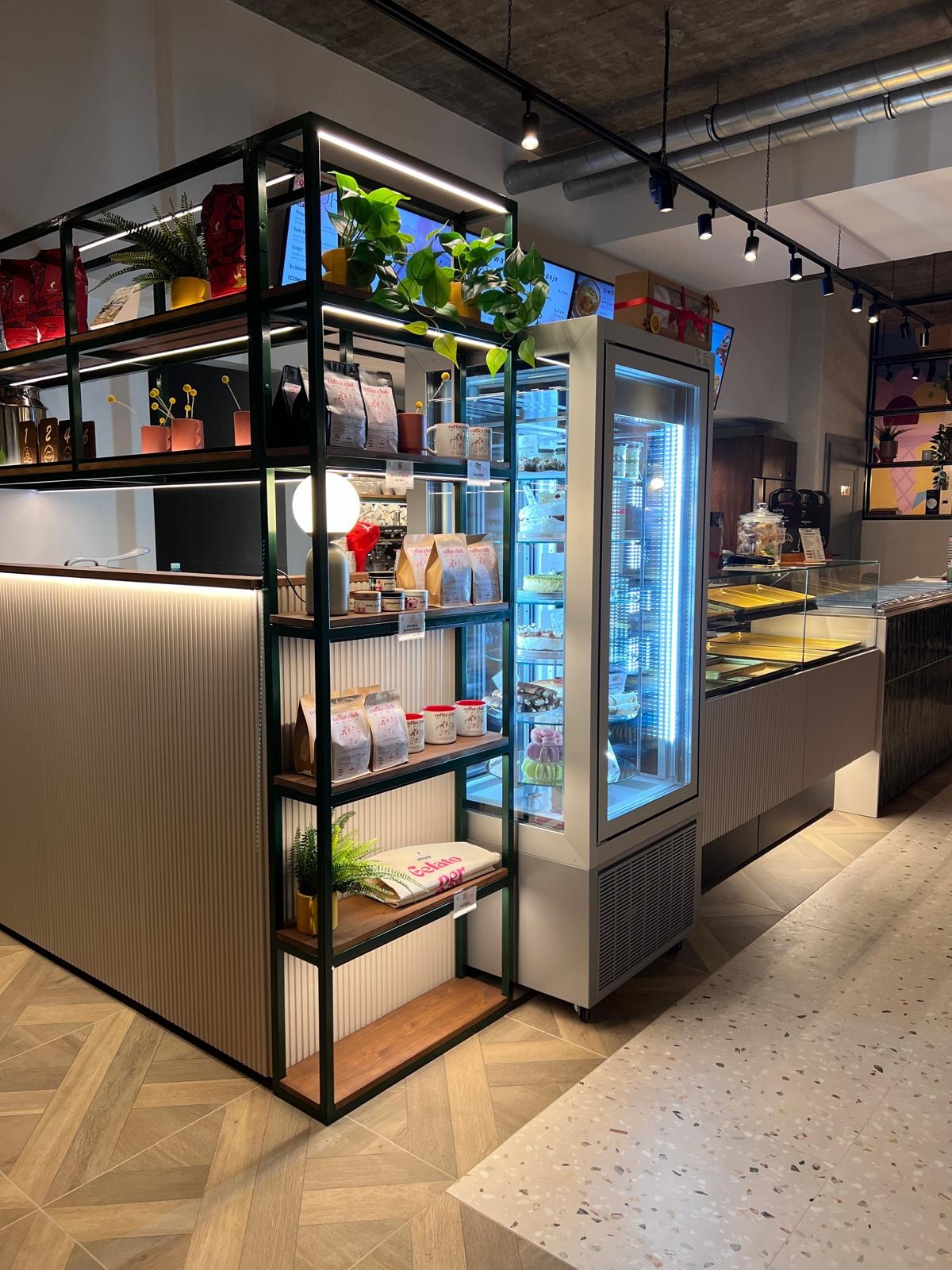 Nowoczesne wnętrze kawiarni z regałem pełnym produktów i roślin, obok chłodnicza witryna z deserami. Parkiet w jodełkę i oświetlenie punktowe.
