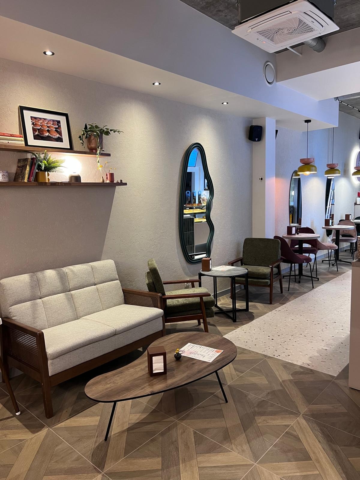 Wnętrze kawiarni z drewnianą podłogą w jodełkę, szarą sofą i stolikiem kawowym. Na ścianie półka z dekoracjami i lustro o nieregularnym kształcie.