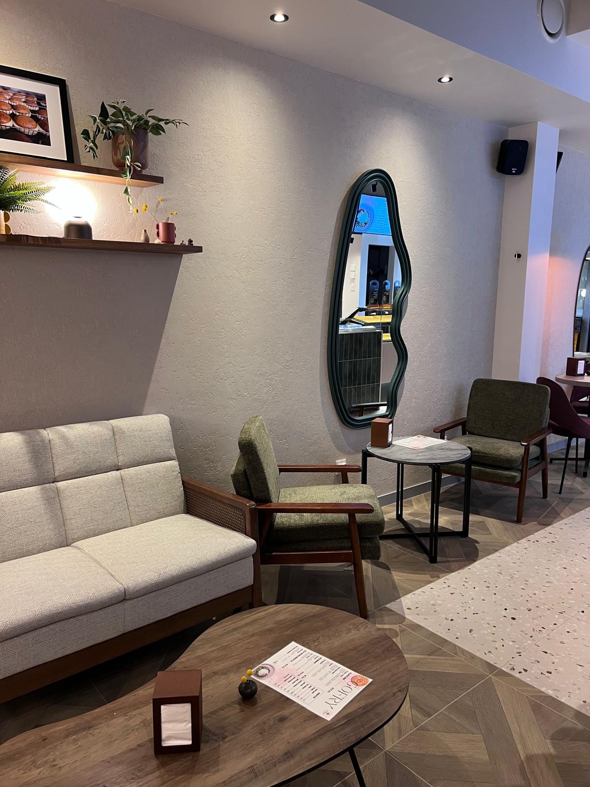 Stylowe wnętrze kawiarni z szarą sofą, zielonymi fotelami i lustrem o nieregularnym kształcie. Na drewnianym stoliku menu. Podłoga z wzorem jodełki i terazzo.