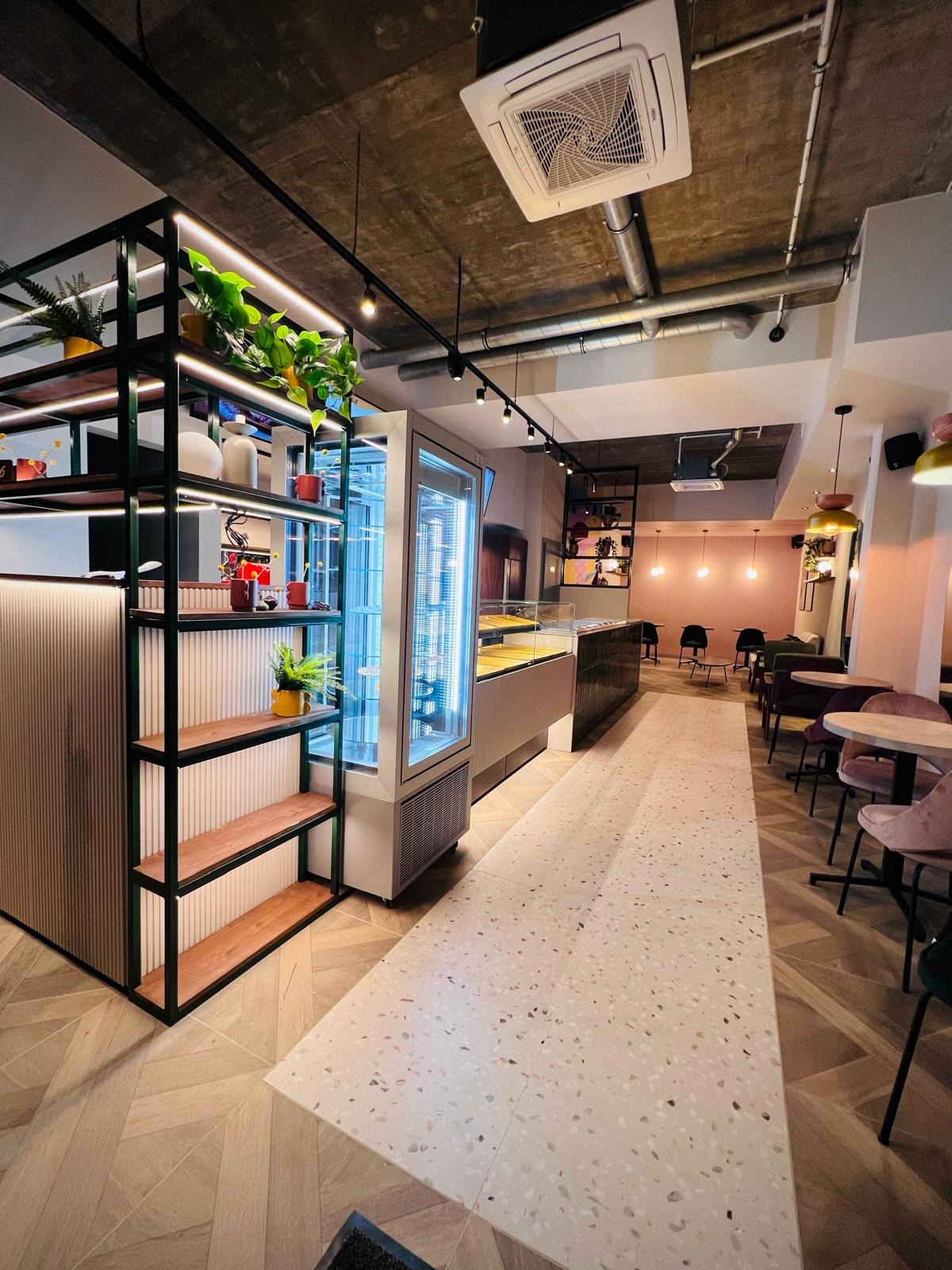 Nowoczesne wnętrze kawiarni z drewnianą podłogą w jodełkę, regałem z roślinami i chłodniczą witryną. Pastelowe ściany i designerskie lampy tworzą przytulną atmosferę.