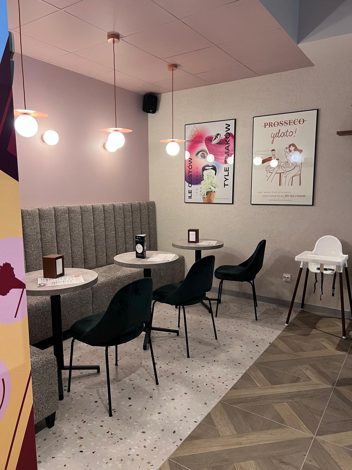 Pastelowe wnętrze kawiarni z szarą tapicerowaną ławą, okrągłymi stolikami, zielonymi krzesłami i plakatami lodów na ścianie. Podłoga w dwóch wzorach.
