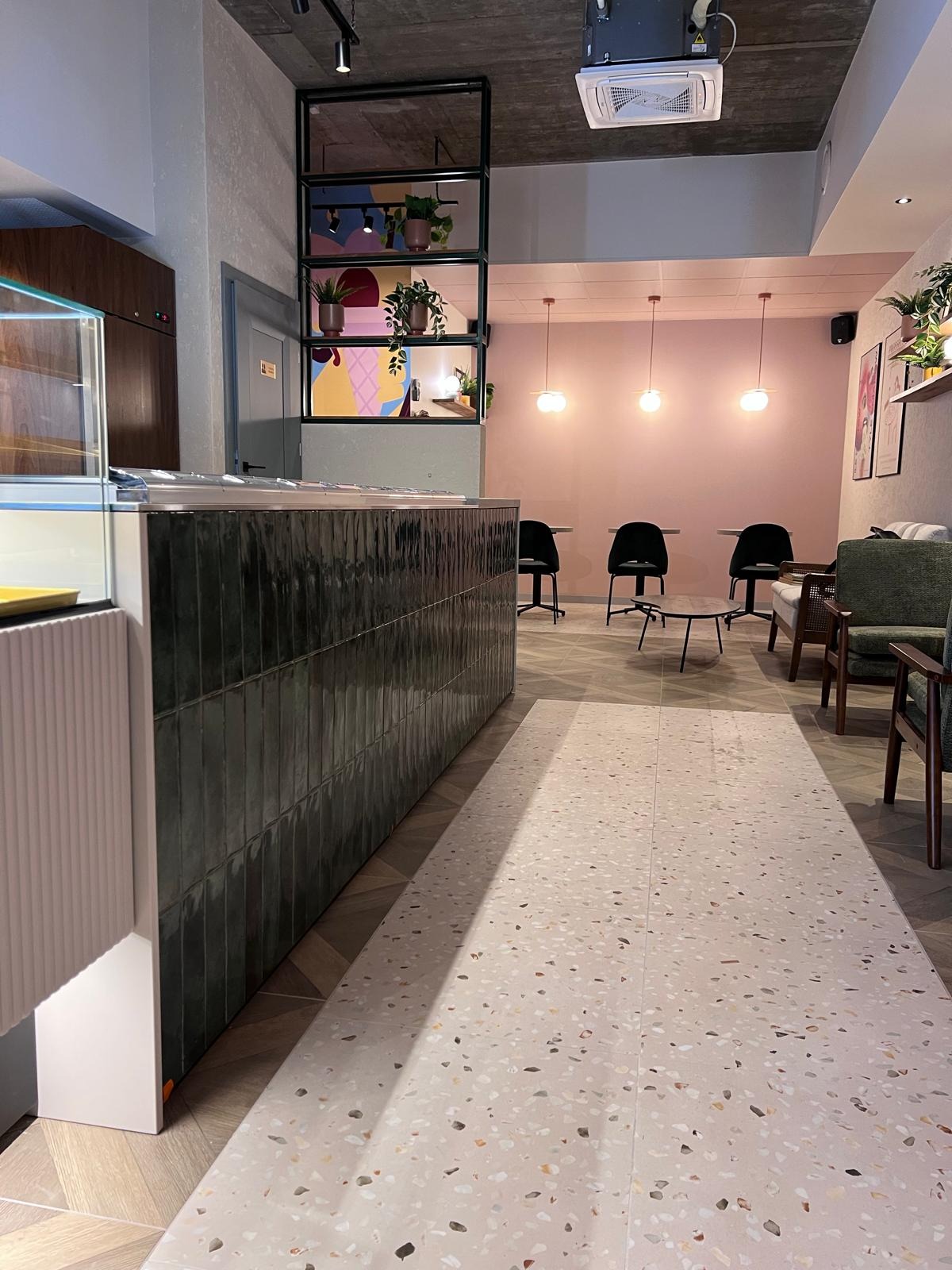 Nowoczesne wnętrze kawiarni z terazzo podłogą i ladą z ciemnej glazury. Pastelowe ściany, minimalistyczne oświetlenie i rośliny w tle tworzą przytulną atmosferę.