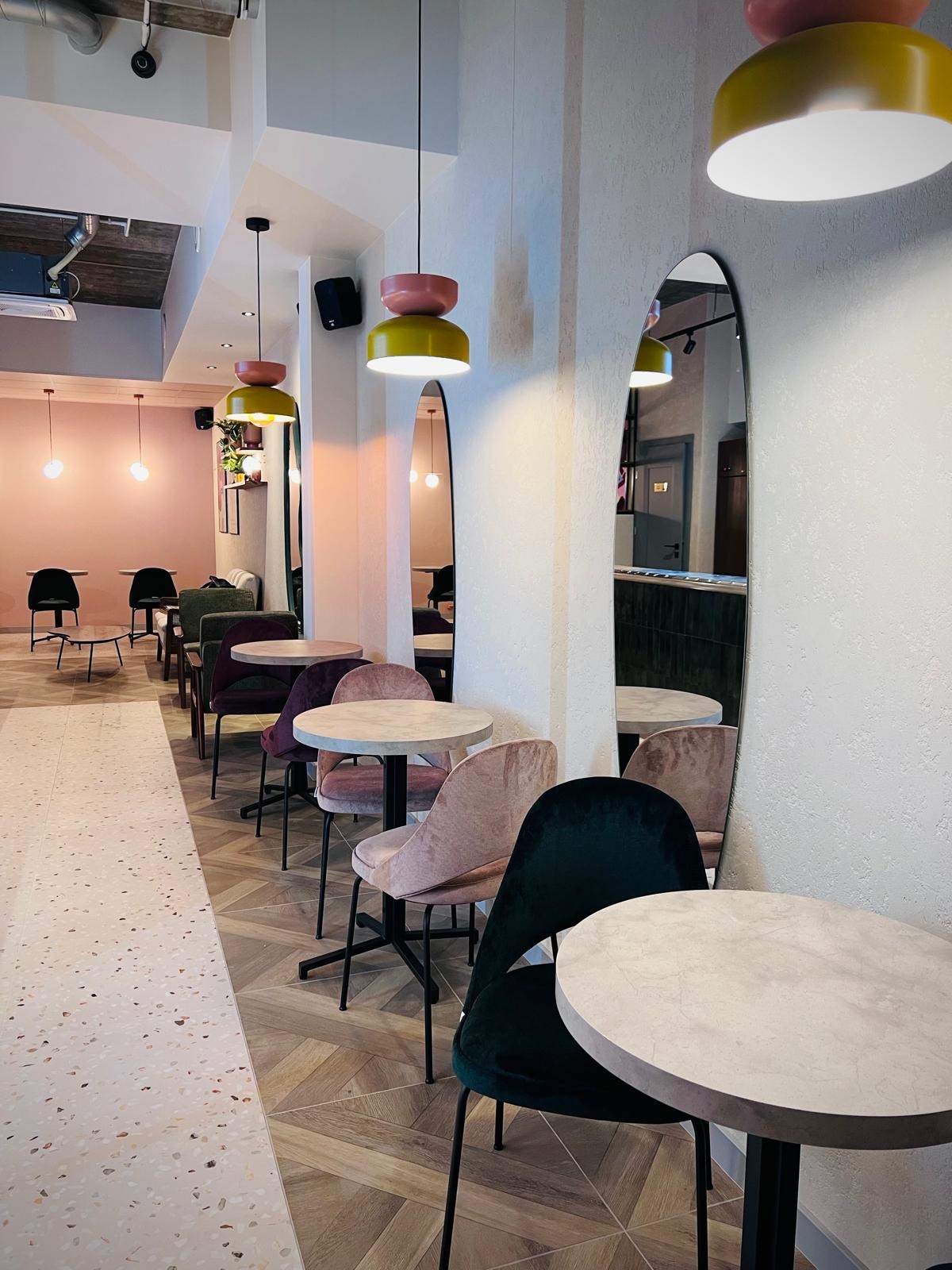 Pastelowe wnętrze kawiarni z rzędem okrągłych stolików i krzeseł w odcieniach różu i zieleni, oświetlone lampami w stylu retro i ozdobione wysokimi lustrami.