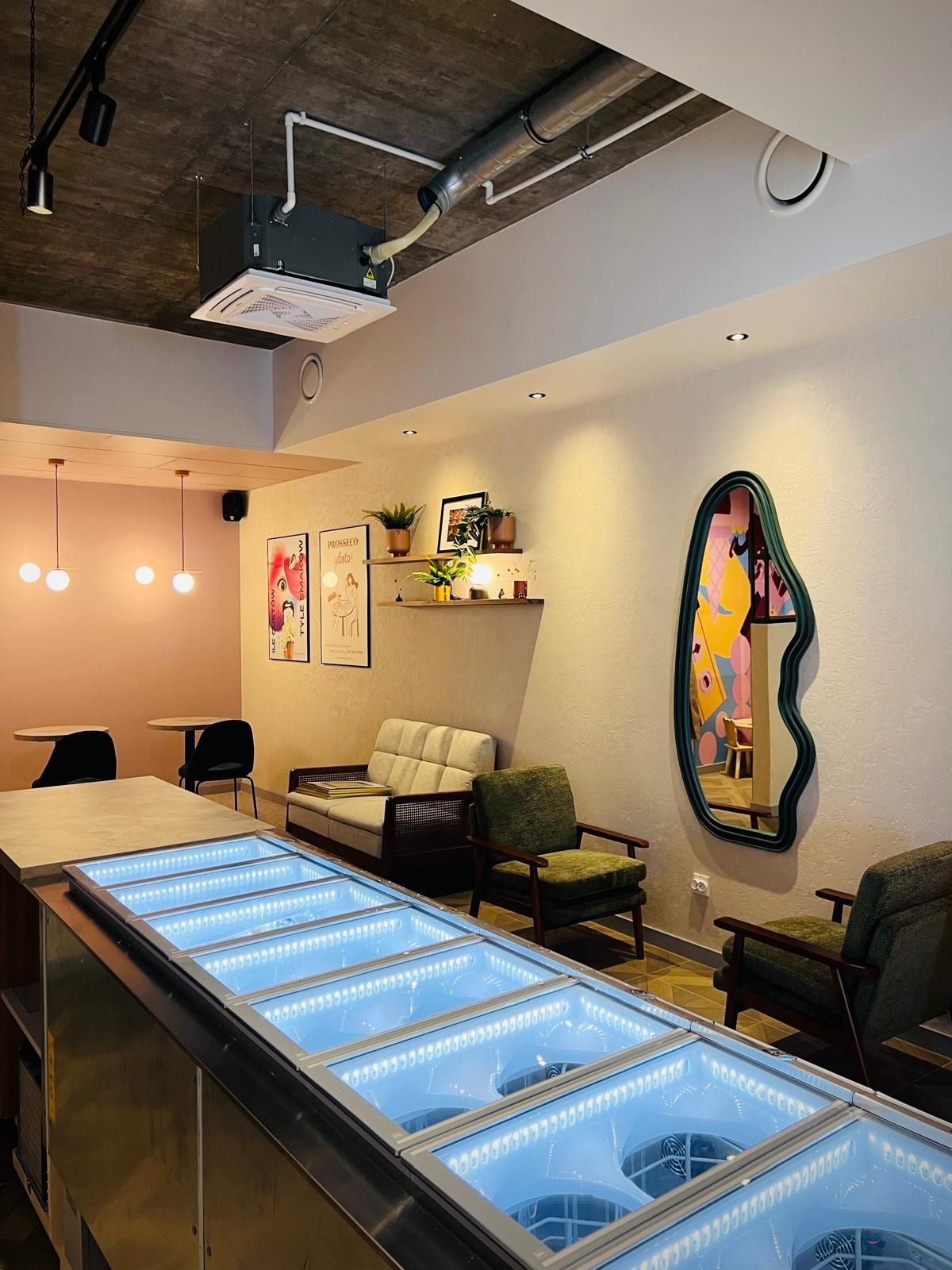 Nowoczesne wnętrze kawiarni z podświetlanym barem, zielonymi fotelami i designerskim lustrem. Widoczny system klimatyzacji pod sufitem. Pastelowe ściany i plakaty.