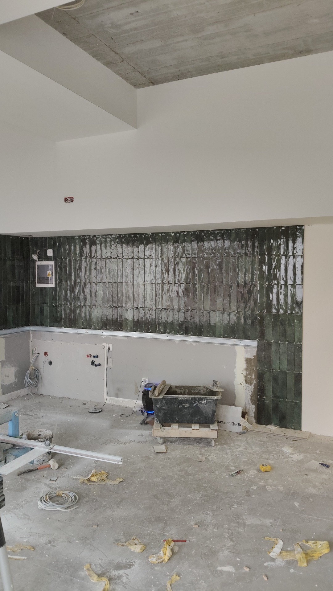 Ściana z połyskującymi, zielonymi płytkami w trakcie montażu. Widoczne narzędzia glazurnicze i surowy betonowy sufit. Częściowo wykończona kuchnia.