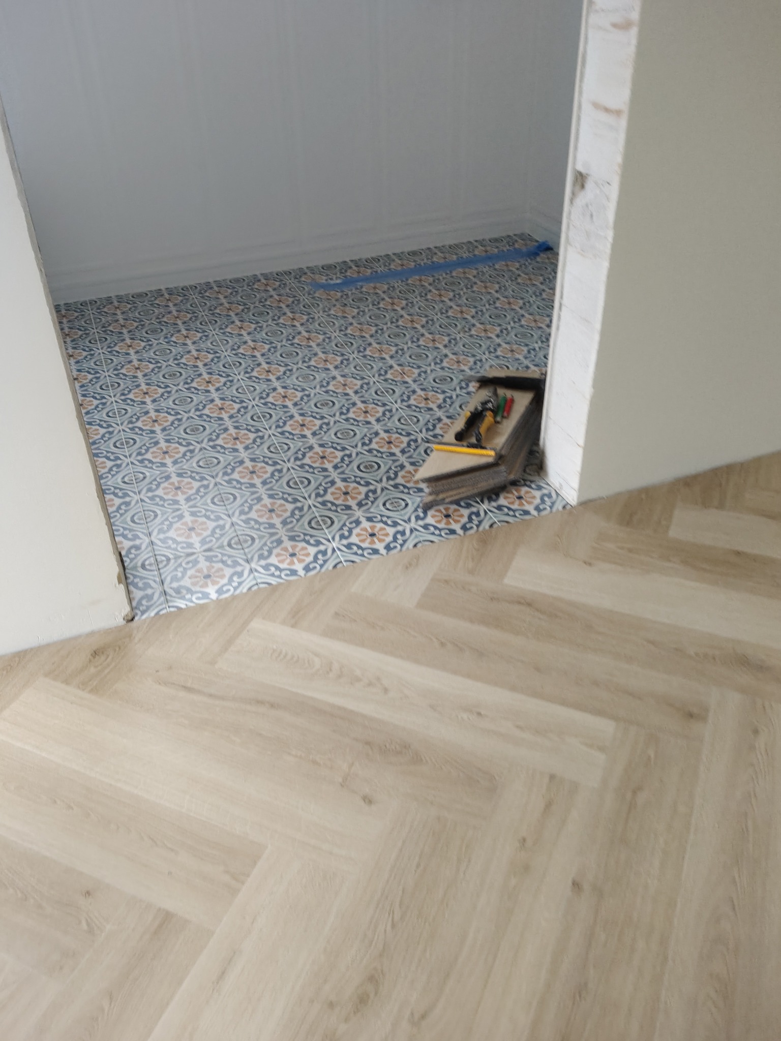 Ułożone płytki wzorzyste w łazience przechodzące w panele podłogowe ułożone w jodełkę. Na płytkach leżą narzędzia glazurnicze.