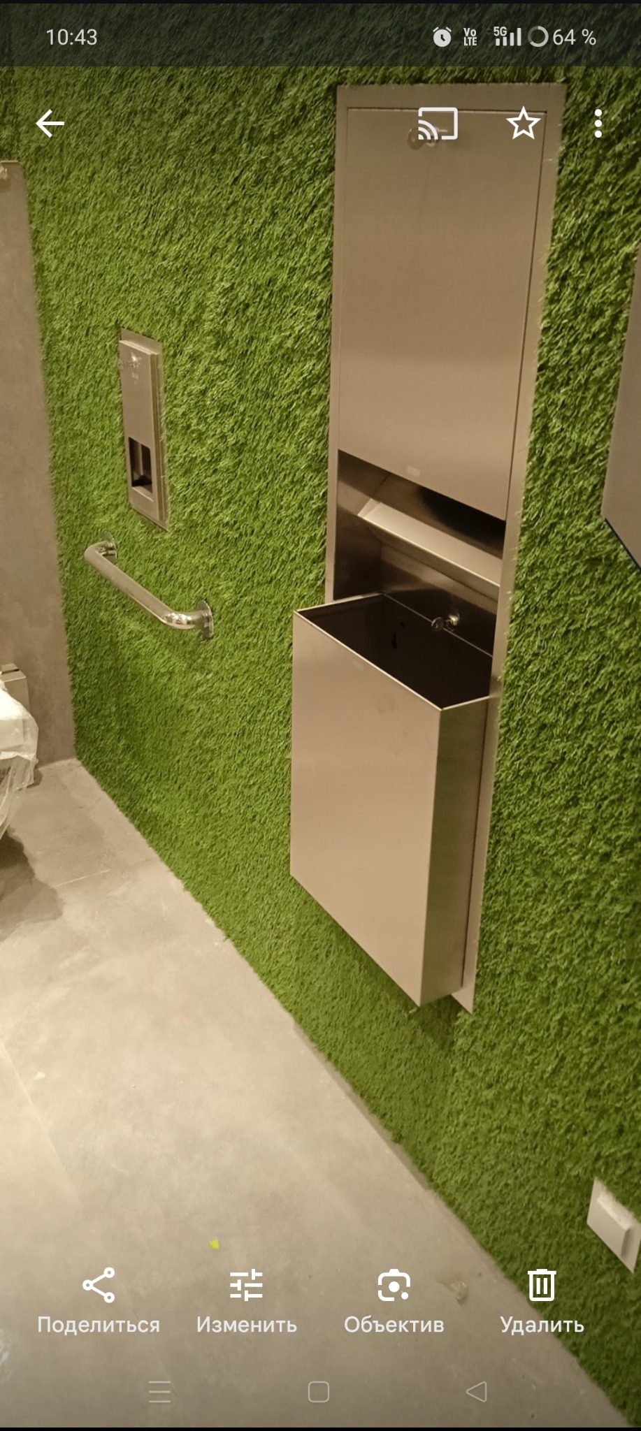 Nowoczesna łazienka z zieloną ścianą imitującą trawę, wyposażona w metalowe akcesoria: podajnik na ręczniki i kosz na śmieci. Dodatkowo zamontowany uchwyt dla niepełnosprawnych.