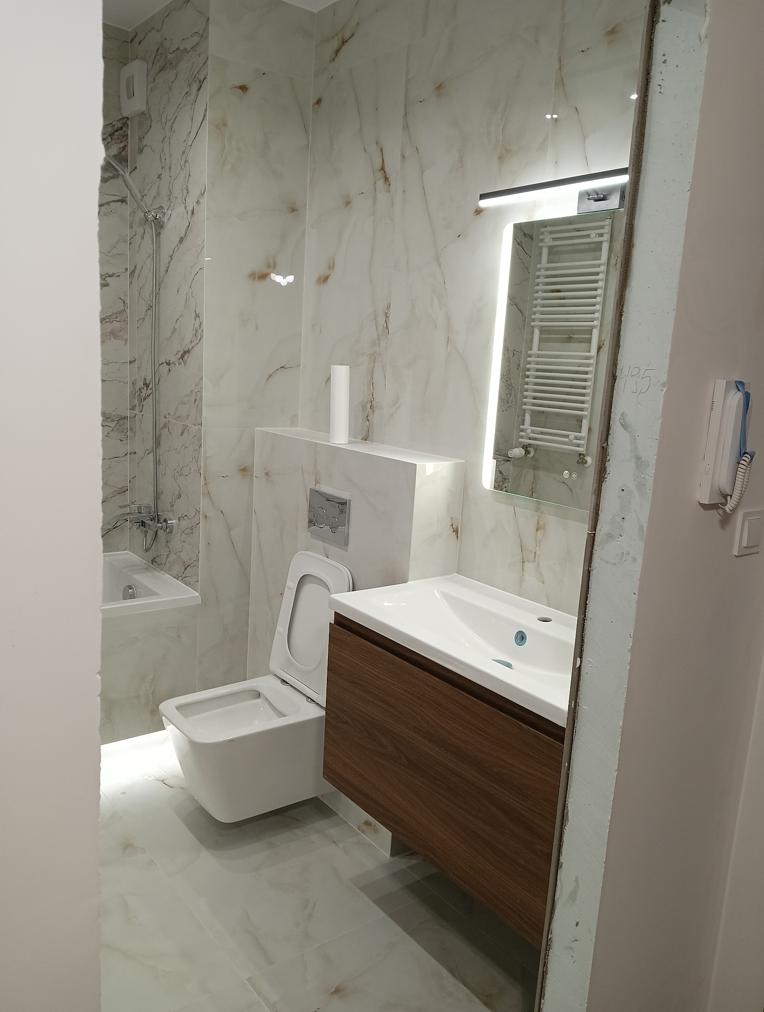 Wykończona łazienka z marmurowymi płytkami, wiszącą toaletą i umywalką z szafką z drewna. Lustro z podświetleniem LED. Widoczny fragment niedokończonej ściany.