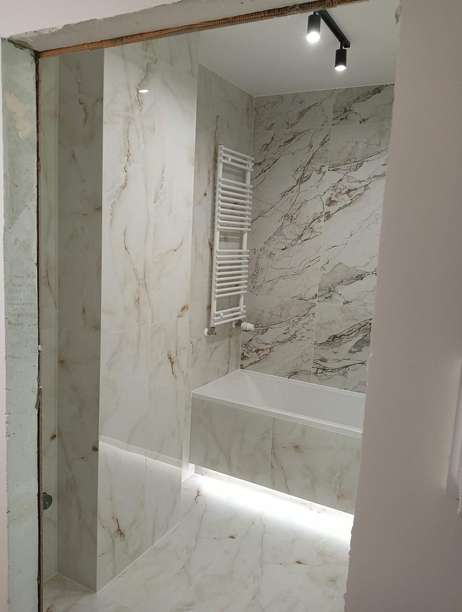 Wykończona łazienka z marmurowymi płytkami, podświetlaną wanną i grzejnikiem drabinkowym. Widok zza framugi, podkreślający efekt końcowy remontu.