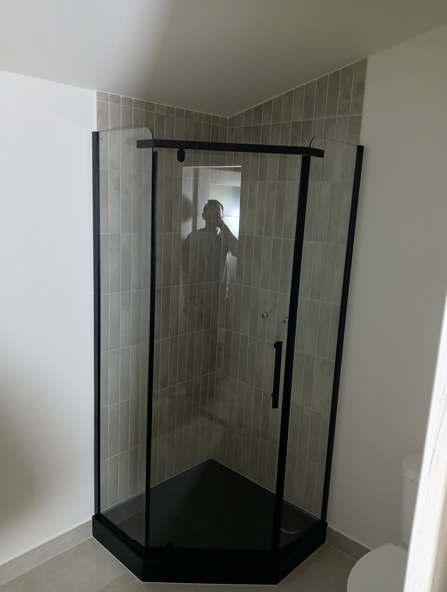 Narożna kabina prysznicowa z czarnymi profilami i szarymi, pionowo ułożonymi płytkami w wyremontowanej łazience. W odbiciu szyby widoczna osoba robiąca zdjęcie.