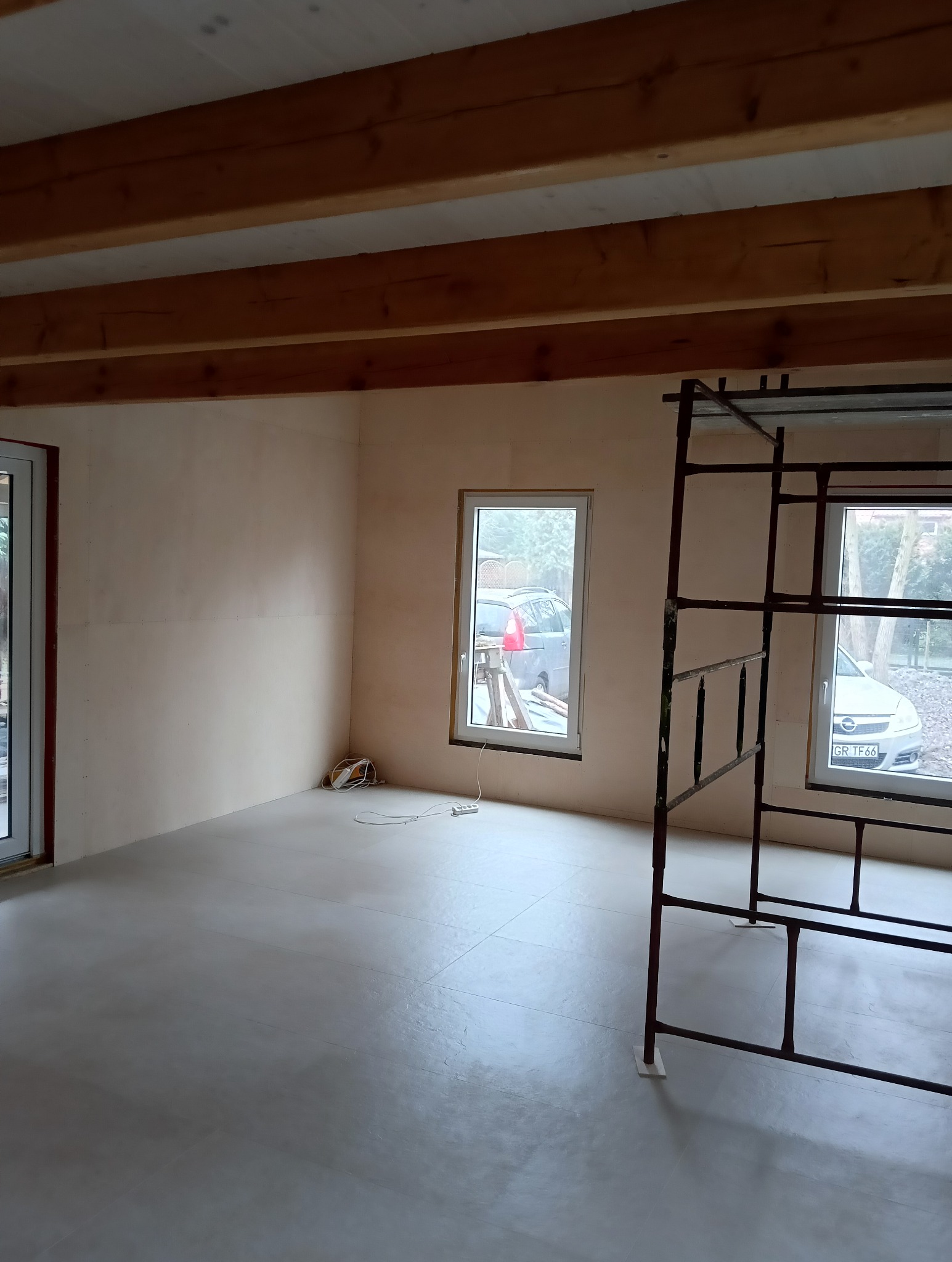 Wnętrze w trakcie prac wykończeniowych: surowe ściany, drewniane belki stropowe, okna z widokiem na zewnątrz, rusztowanie w rogu. Widoczne kable i gładka podłoga.