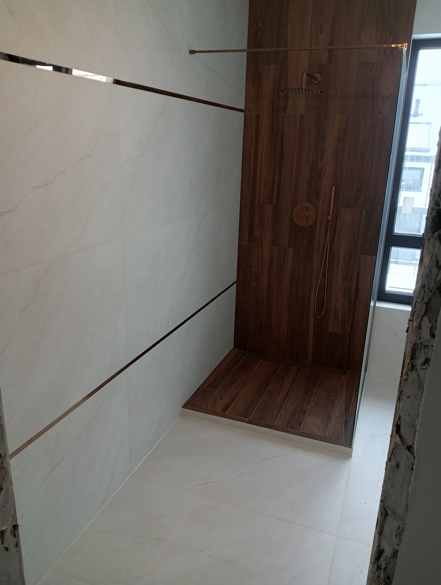 Nowoczesna kabina prysznicowa z drewnopodobnymi płytkami i białymi płytkami z dekoracyjnymi listwami w kolorze miedzi. Widoczna armatura w odcieniach miedzi.