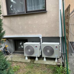 Kosmala - Dwa, zamontowane na zewnątrz, klimatyzatory Gree Inverter, stojące na białych nóżkach przy ścianie budynku z oknem i zielenią. Widoczne rury odprowadzające.