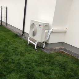 Kosmala - Biała jednostka zewnętrzna klimatyzacji Gree, zamontowana na wspornikach przy ścianie budynku z trawnikiem. Widoczne rury w białej osłonie.