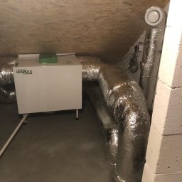 Kosmala - Biała jednostka rekuperacji WANAS z srebrnymi, izolowanymi przewodami wentylacyjnymi w szarej przestrzeni poddasza z drewnianym wykończeniem stropu.