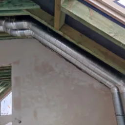 Dwa srebrne przewody wentylacyjne, owinięte taśmą, zamontowane pod drewnianą konstrukcją dachu na poddaszu w trakcie prac wykończeniowych.