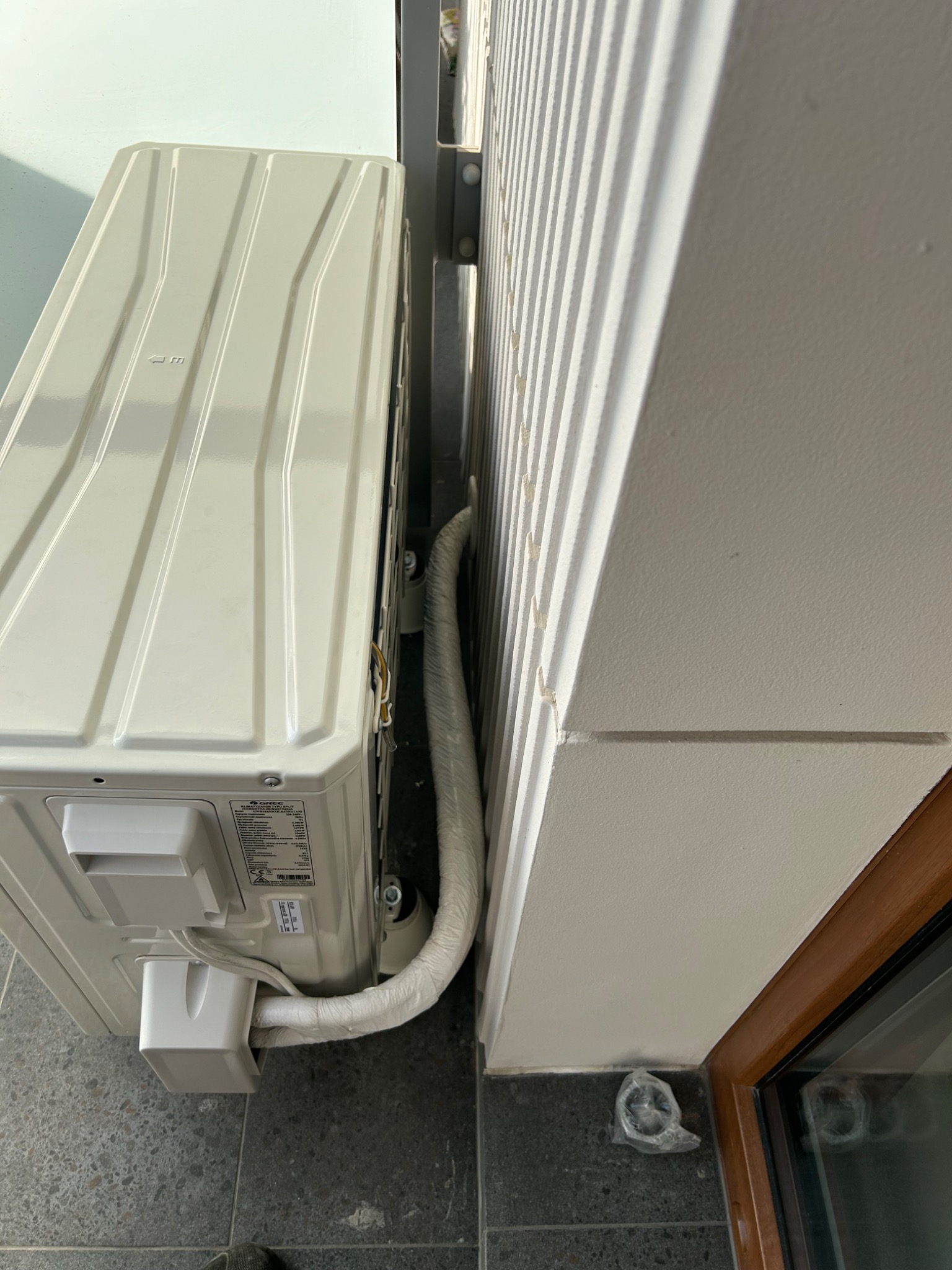 Zamontowana jednostka zewnętrzna klimatyzacji marki Gree, z widocznymi przewodami i elementami instalacji, umieszczona na szarej, kafelkowej podłodze przy ścianie.