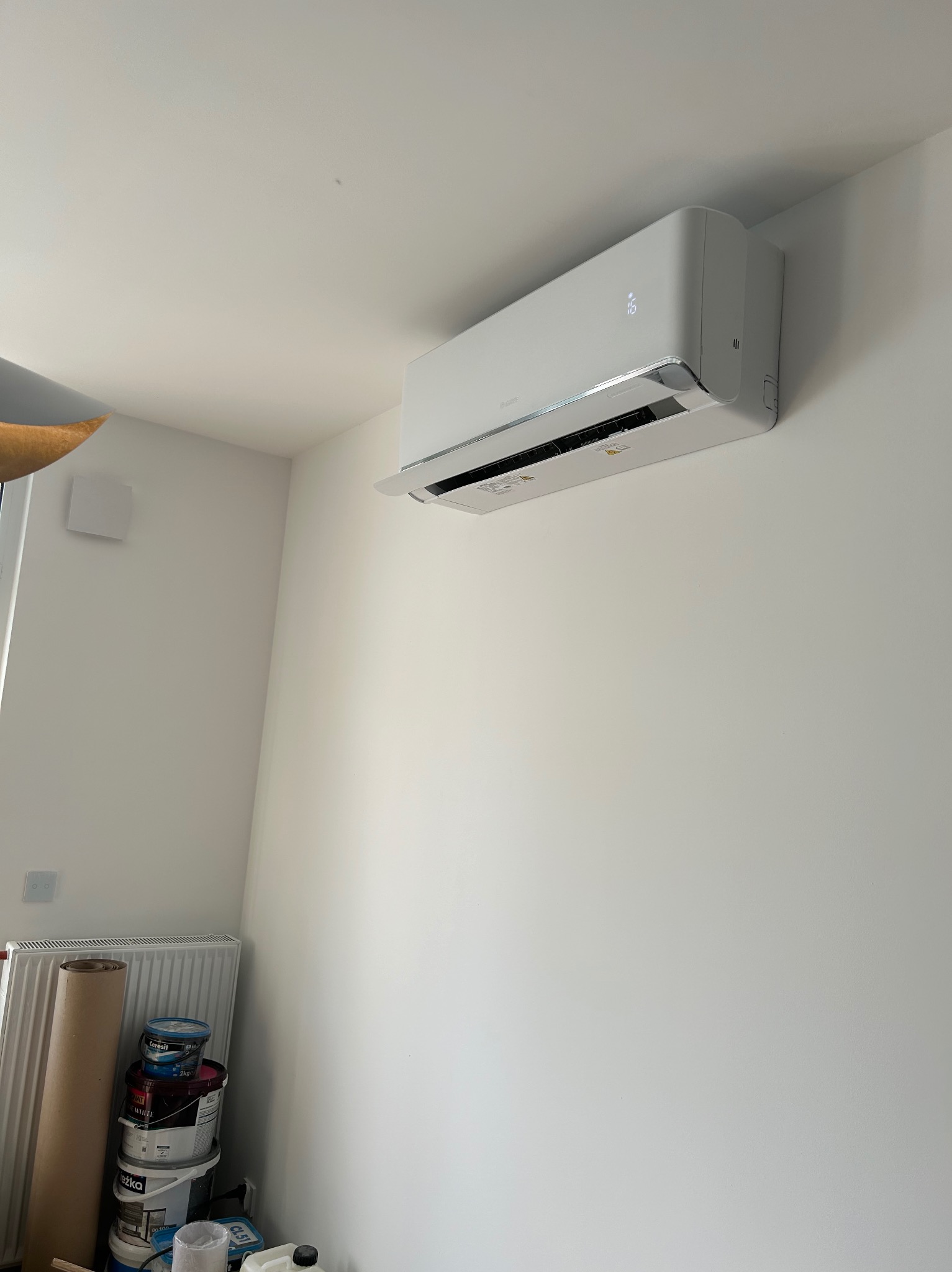Biały klimatyzator ścienny zamontowany w jasnym pomieszczeniu, widoczne farby malarskie i grzejnik. Minimalistyczny design, świeżo po instalacji.