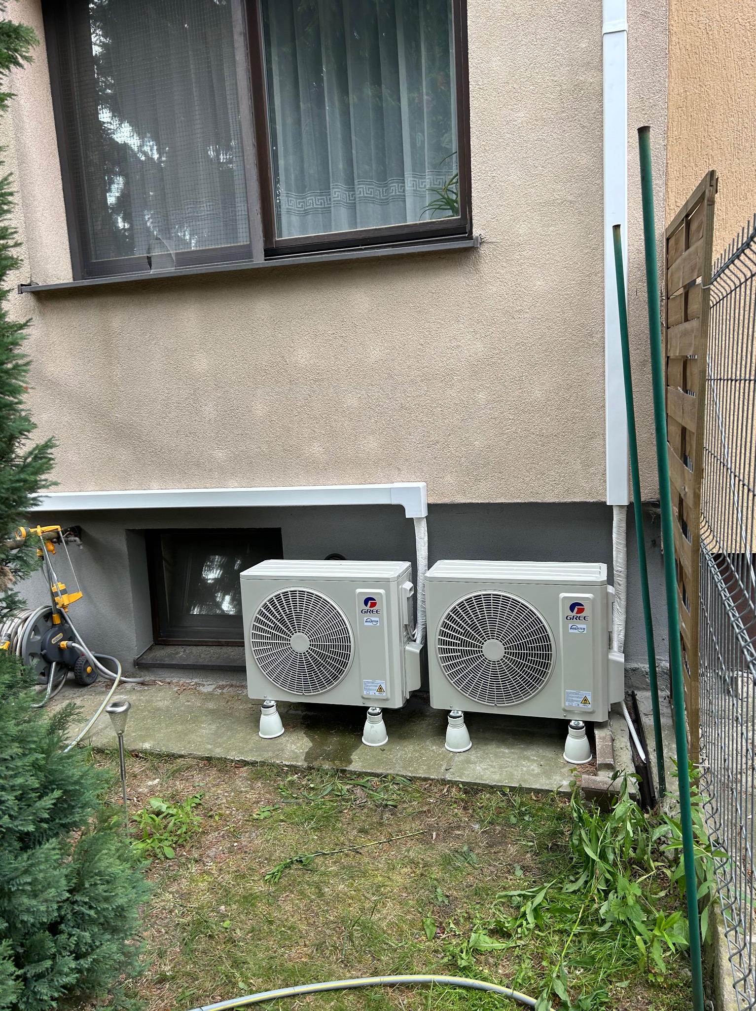 Dwa, zamontowane na zewnątrz, klimatyzatory Gree Inverter, stojące na białych nóżkach przy ścianie budynku z oknem i zielenią. Widoczne rury odprowadzające.