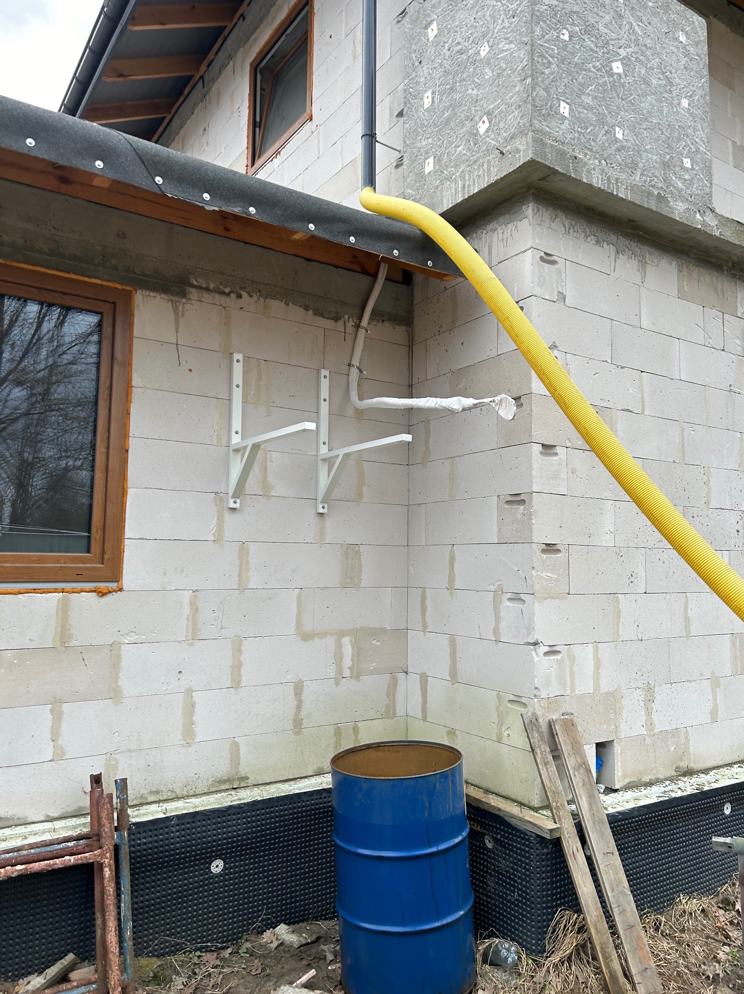 Ściana budynku w trakcie budowy z zamontowanymi wspornikami, żółtą rurą wentylacyjną i niebieską beczką. Widoczne detale montażu i materiały budowlane.