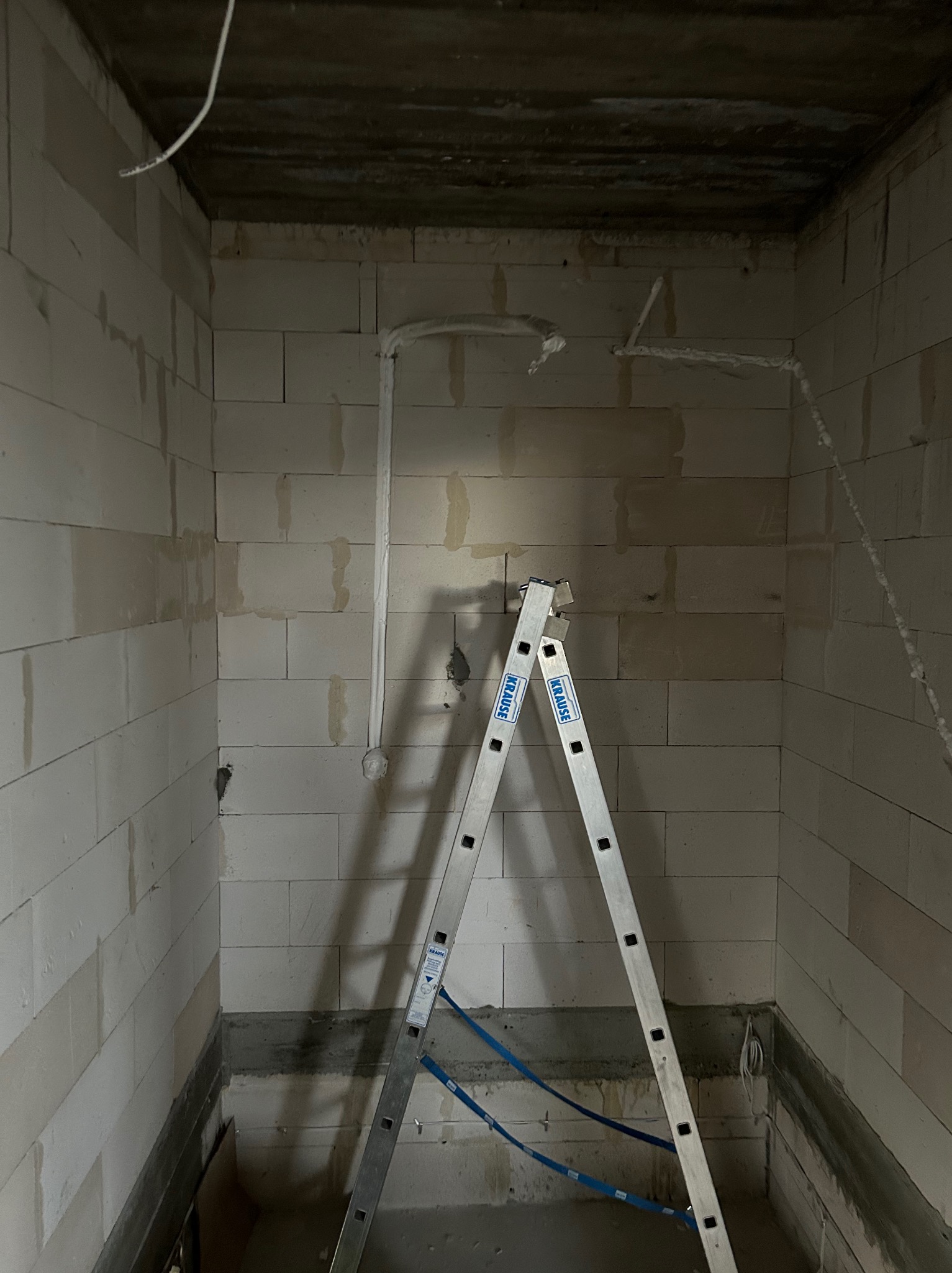 Wewnętrzny kadr z surowymi ścianami z betonu komórkowego, instalacją wentylacyjną pod stropem i rozstawioną drabiną Krause. Etap montażu instalacji.