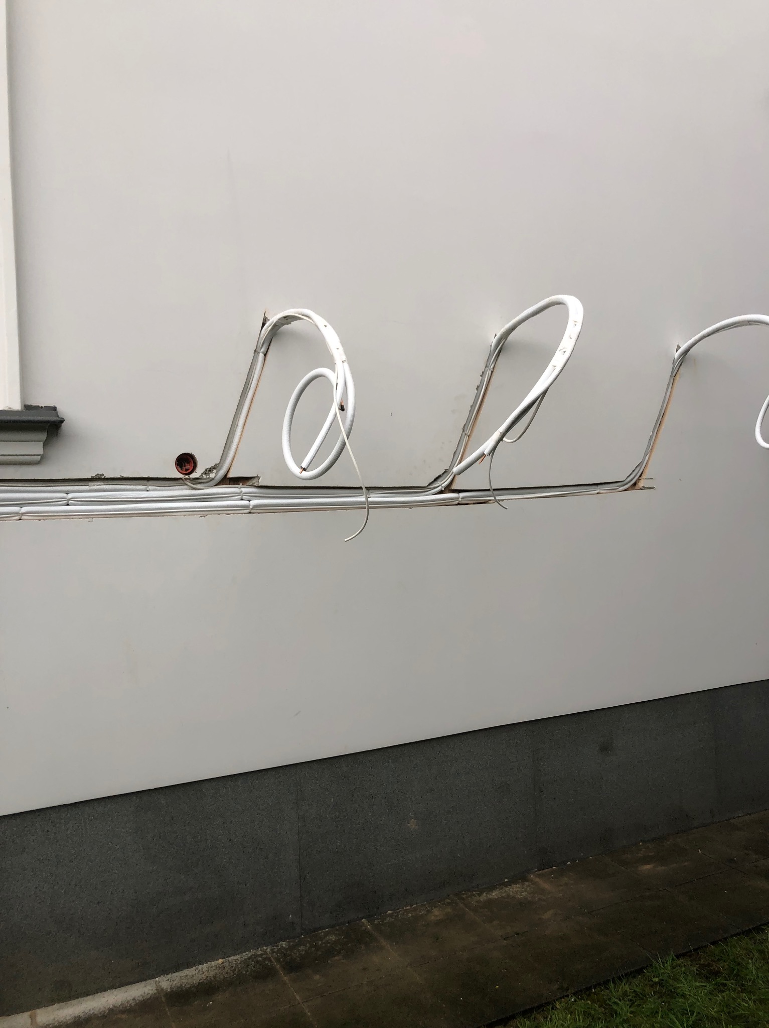 Białe, zaizolowane rury instalacyjne, prowadzone po zewnętrznej ścianie budynku, z widocznymi otworami w ścianie i elementami mocującymi.