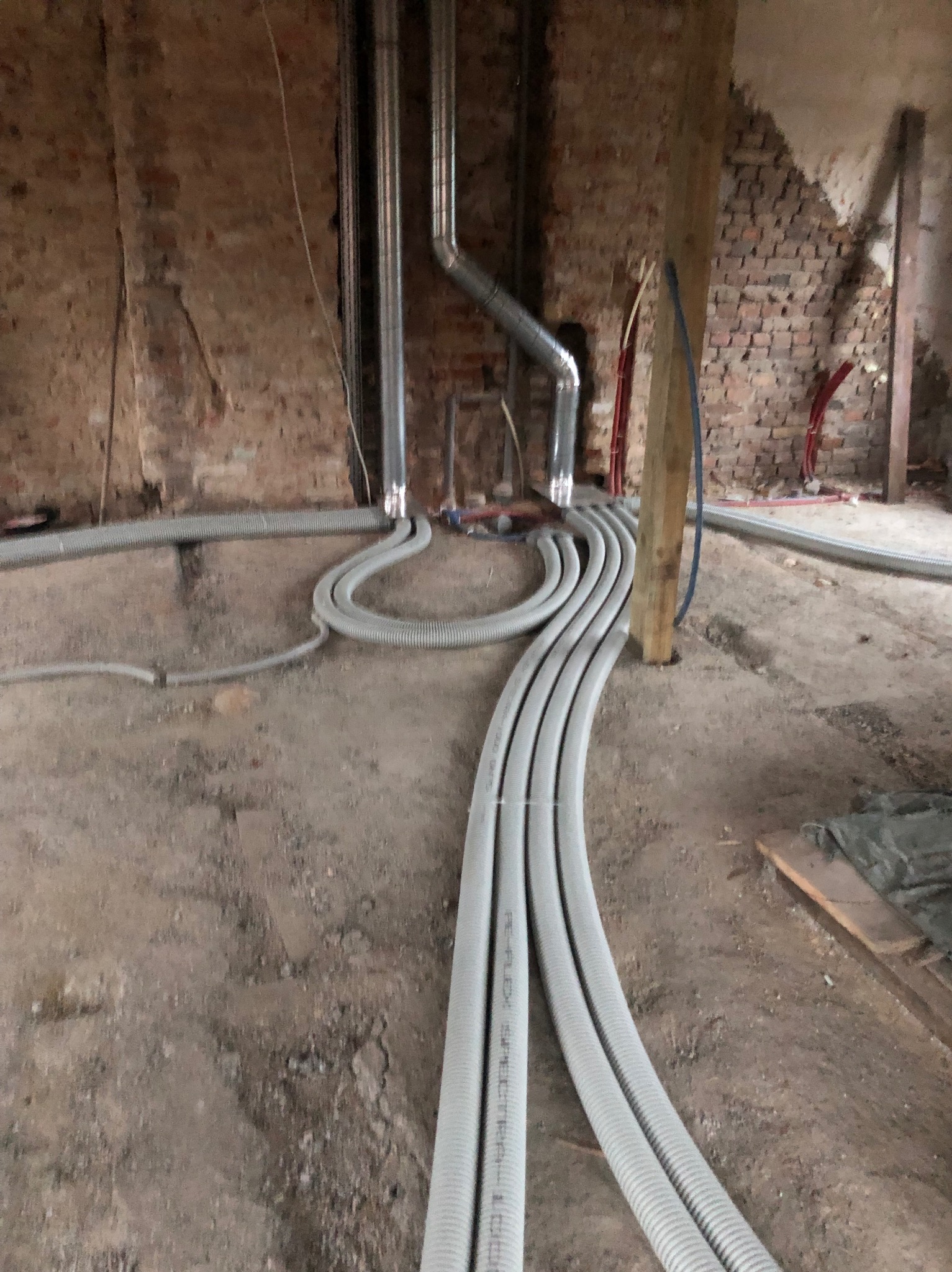 Szare rury wentylacyjne ułożone na podłodze w surowym wnętrzu z ceglanymi ścianami i drewnianymi elementami konstrukcyjnymi. Widoczne pionowe rury i instalacje.