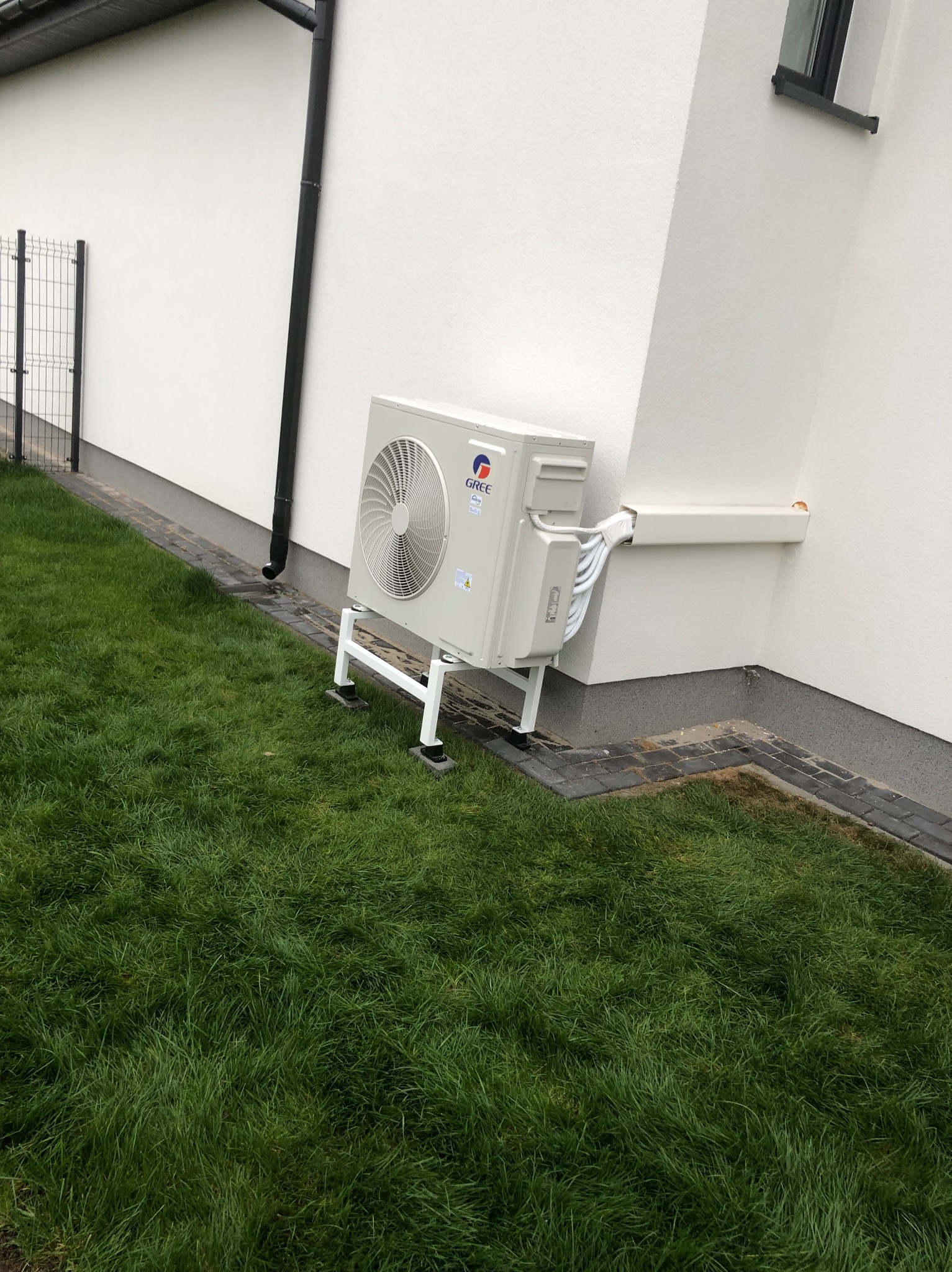 Biała jednostka zewnętrzna klimatyzacji Gree, zamontowana na wspornikach przy ścianie budynku z trawnikiem. Widoczne rury w białej osłonie.
