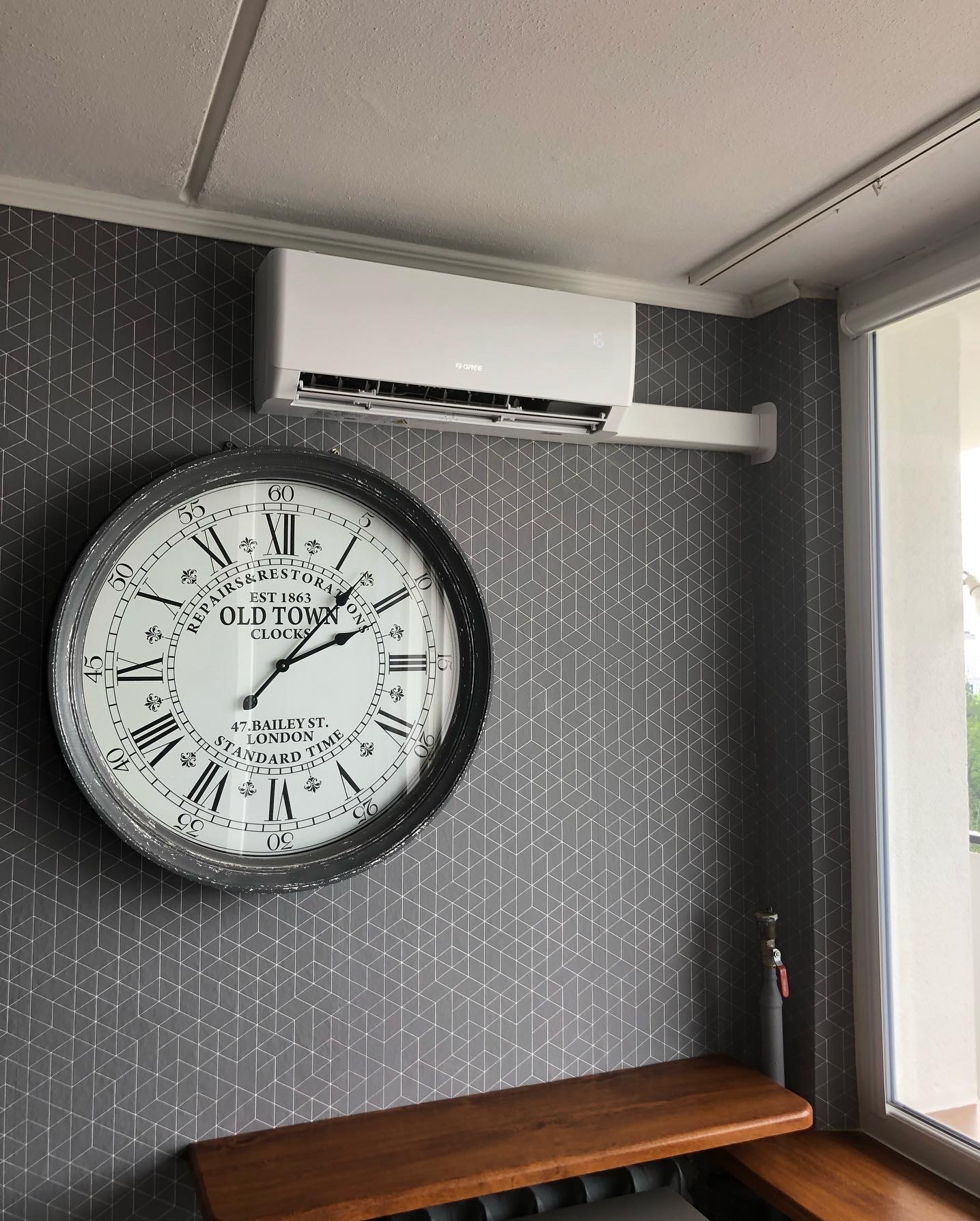 Biały klimatyzator Gree na ścianie z geometrycznym wzorem, obok dekoracyjny zegar w Sulejówku. Widoczny fragment okna i drewniany parapet.