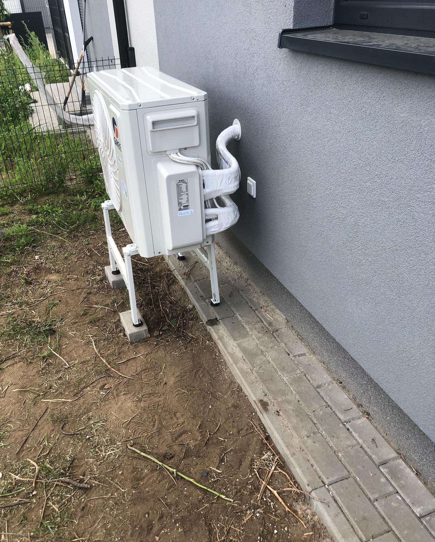 Biała jednostka zewnętrzna klimatyzacji Gree zamontowana na metalowym stelażu przy szarej ścianie budynku. Rury owinięte izolacją.