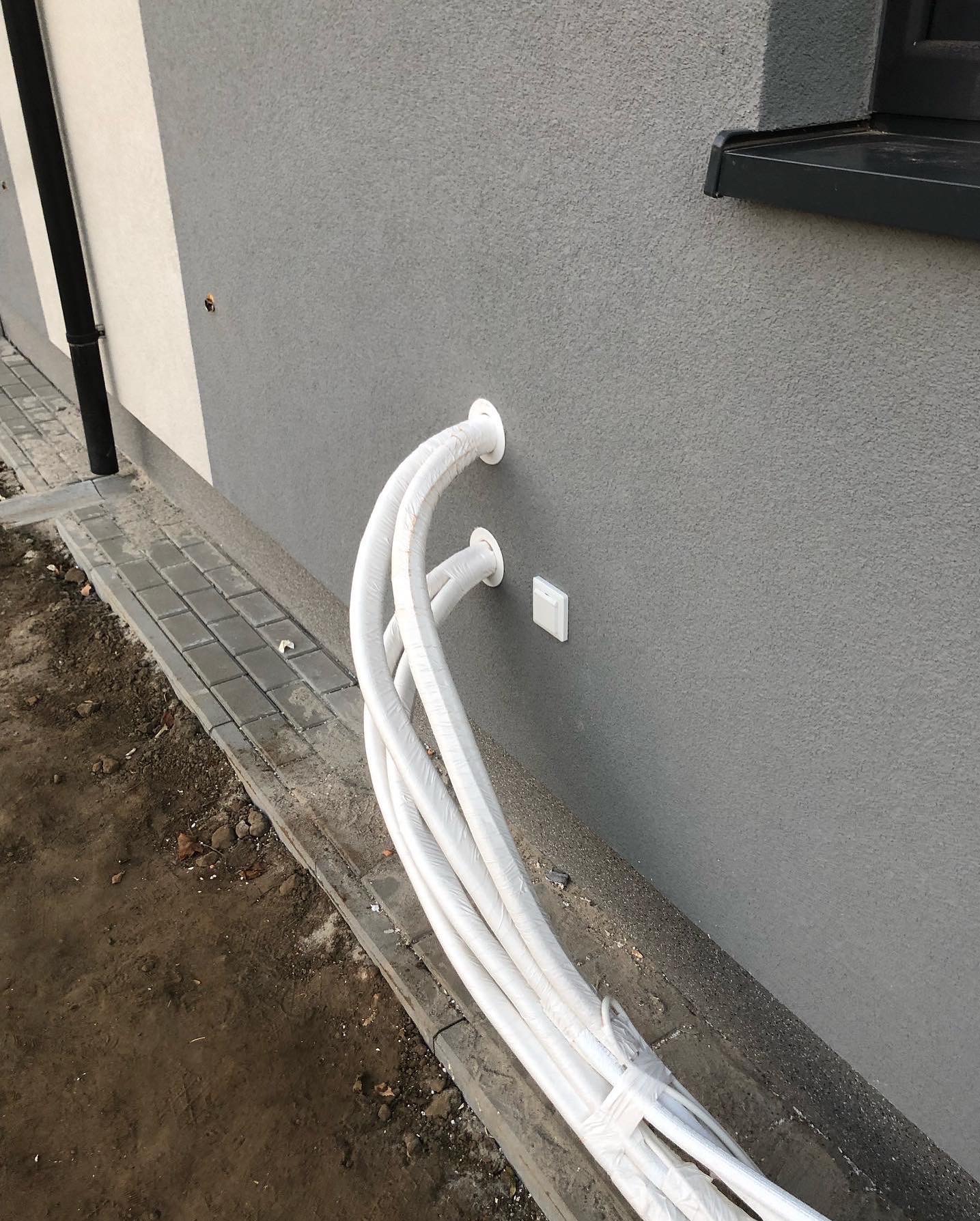 Białe rury w izolacji, wystające ze ściany budynku z szarą elewacją, prawdopodobnie instalacja klimatyzacji lub wentylacji przed uruchomieniem.
