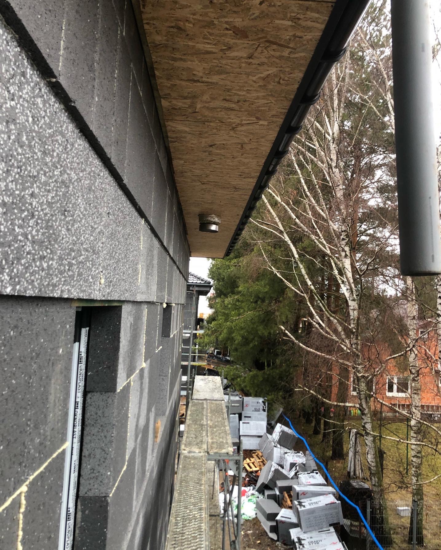 Montaż rekuperacji: Szare bloczki budynku z widoczną instalacją wentylacyjną pod okapem. Na pierwszym planie rusztowanie i materiały budowlane.