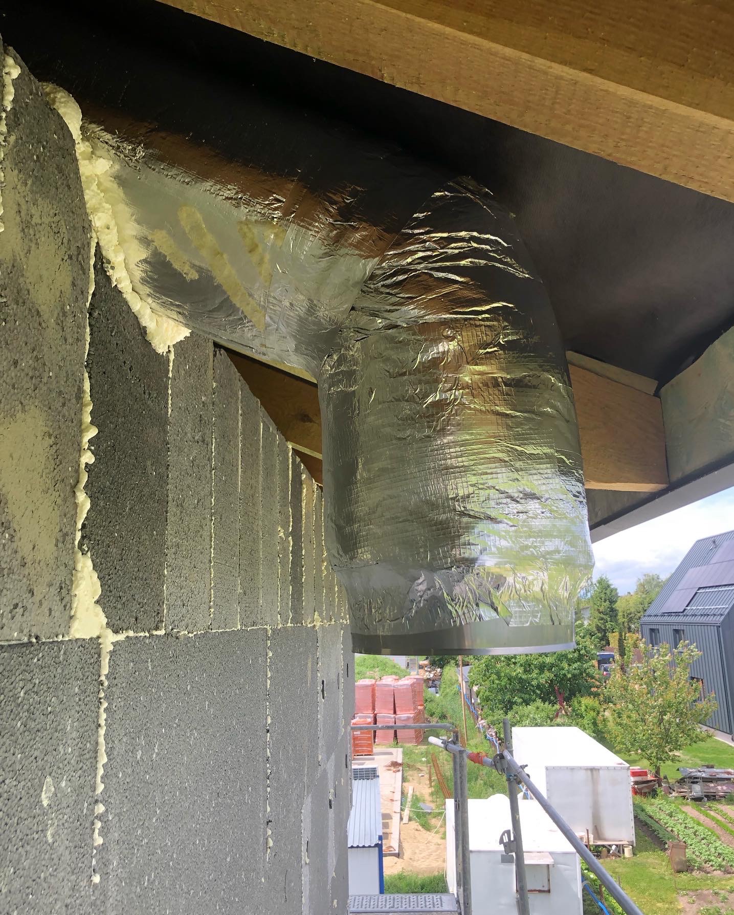 Ocieplony, aluminiową folią kanał wentylacyjny, zamontowany w nowym budynku z szarych bloczków, uszczelniony pianką. Widok na zielone otoczenie i budynki.