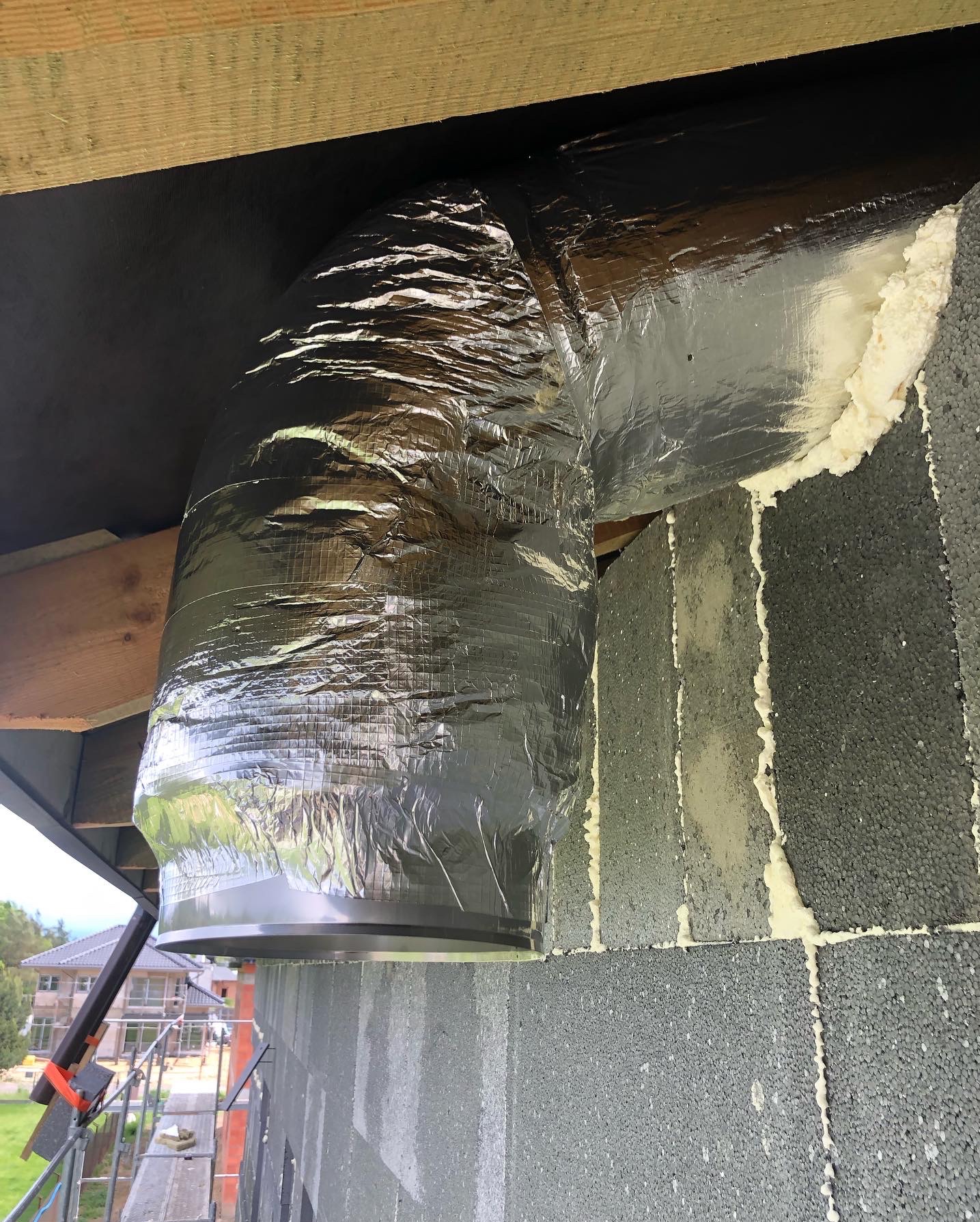 Srebrny, zaizolowany przewód wentylacyjny zamontowany w szarej ścianie z pianką montażową, widoczny na tle konstrukcji budynku.