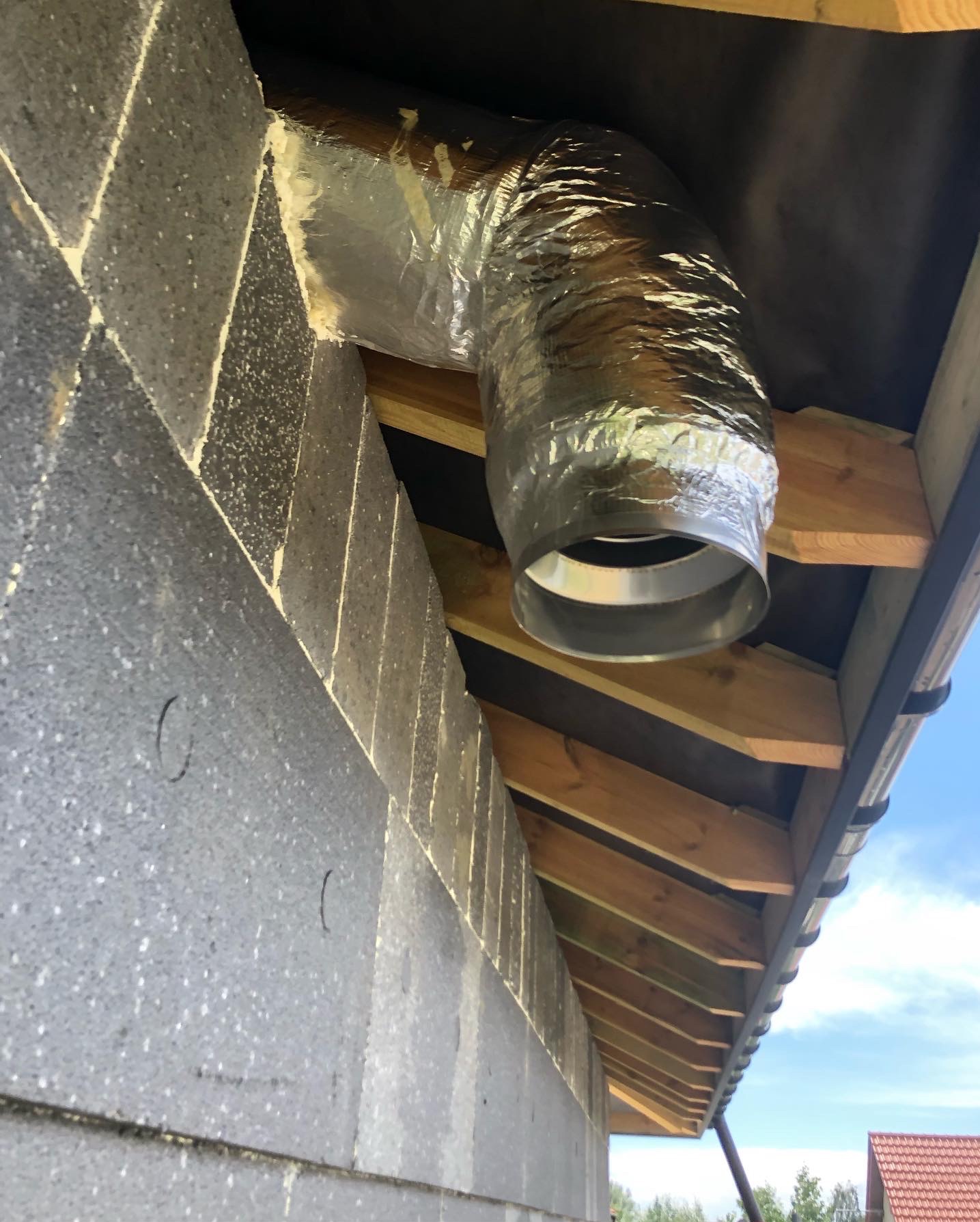 Sztywny, srebrny przewód wentylacyjny pod kątem 90 stopni, przymocowany do ściany z szarego betonu komórkowego i drewnianej konstrukcji dachu.