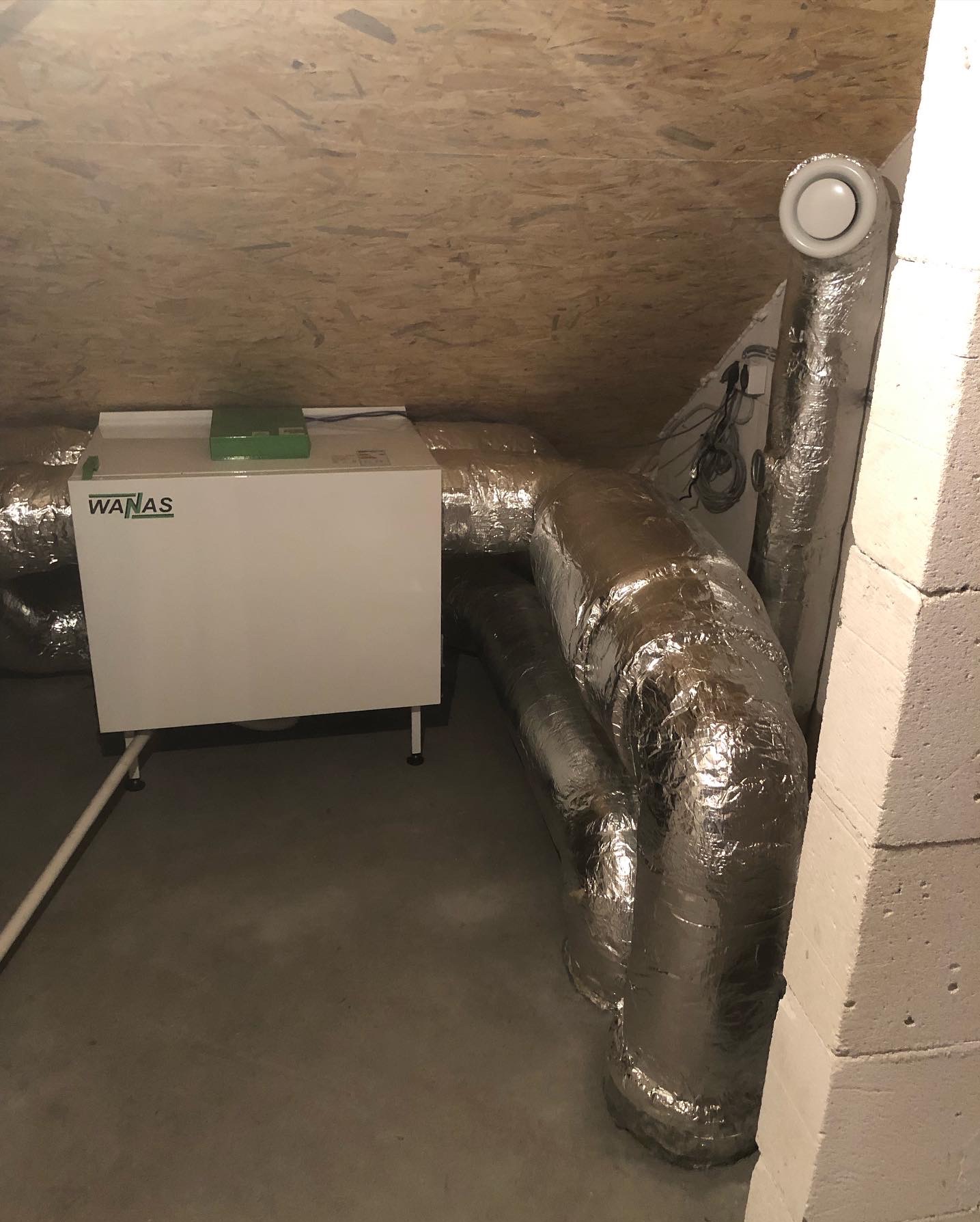 Biała jednostka rekuperacji WANAS z srebrnymi, izolowanymi przewodami wentylacyjnymi w szarej przestrzeni poddasza z drewnianym wykończeniem stropu.