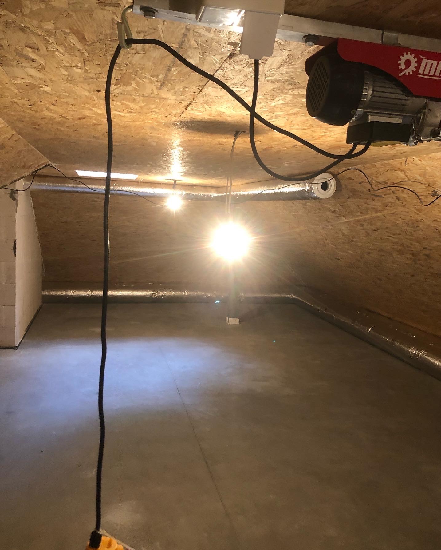 Wykończone poddasze z widocznymi rurami wentylacyjnymi pod drewnianym stropem. Na suficie zainstalowany elektryczny podnośnik. Jasne światło.