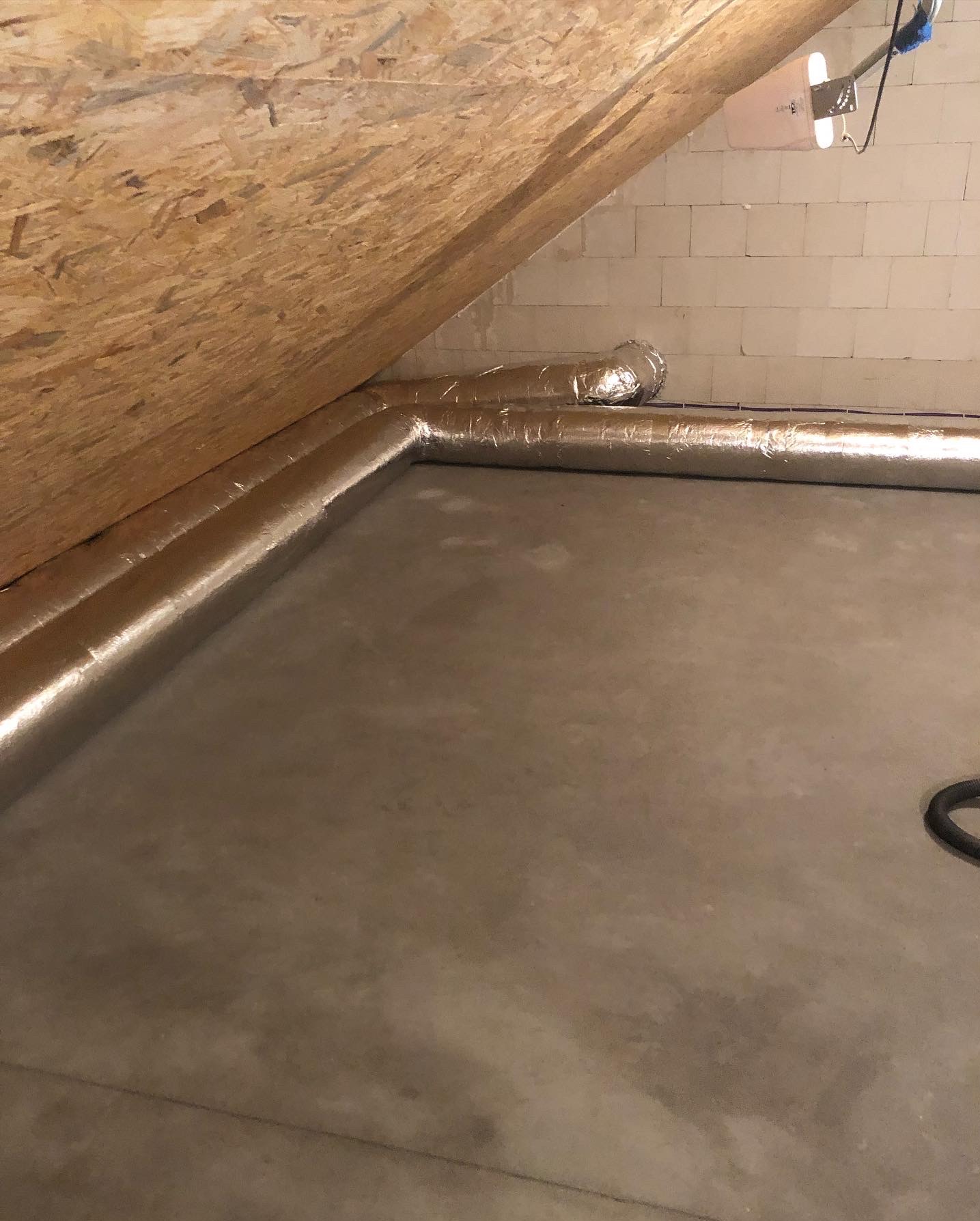 Szczegółowe ujęcie izolowanych kanałów wentylacyjnych w przestrzeni poddasza, z widoczną ścianą z bloczków i drewnianą konstrukcją dachu.