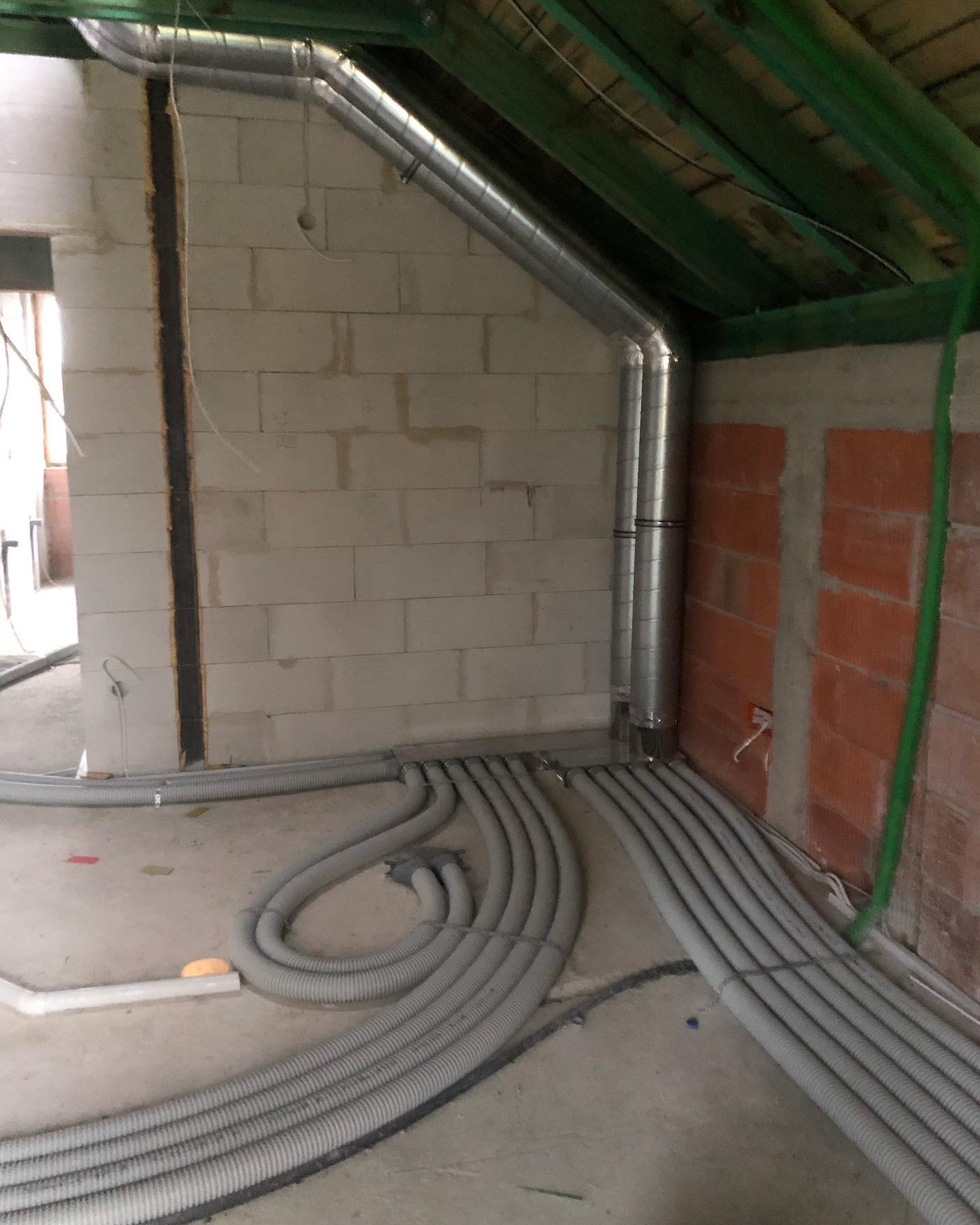 Szare, elastyczne rury wentylacyjne ułożone na podłodze surowego pomieszczenia z ceglanymi i betonowymi ścianami. Metalowe rury pionowo w rogu.