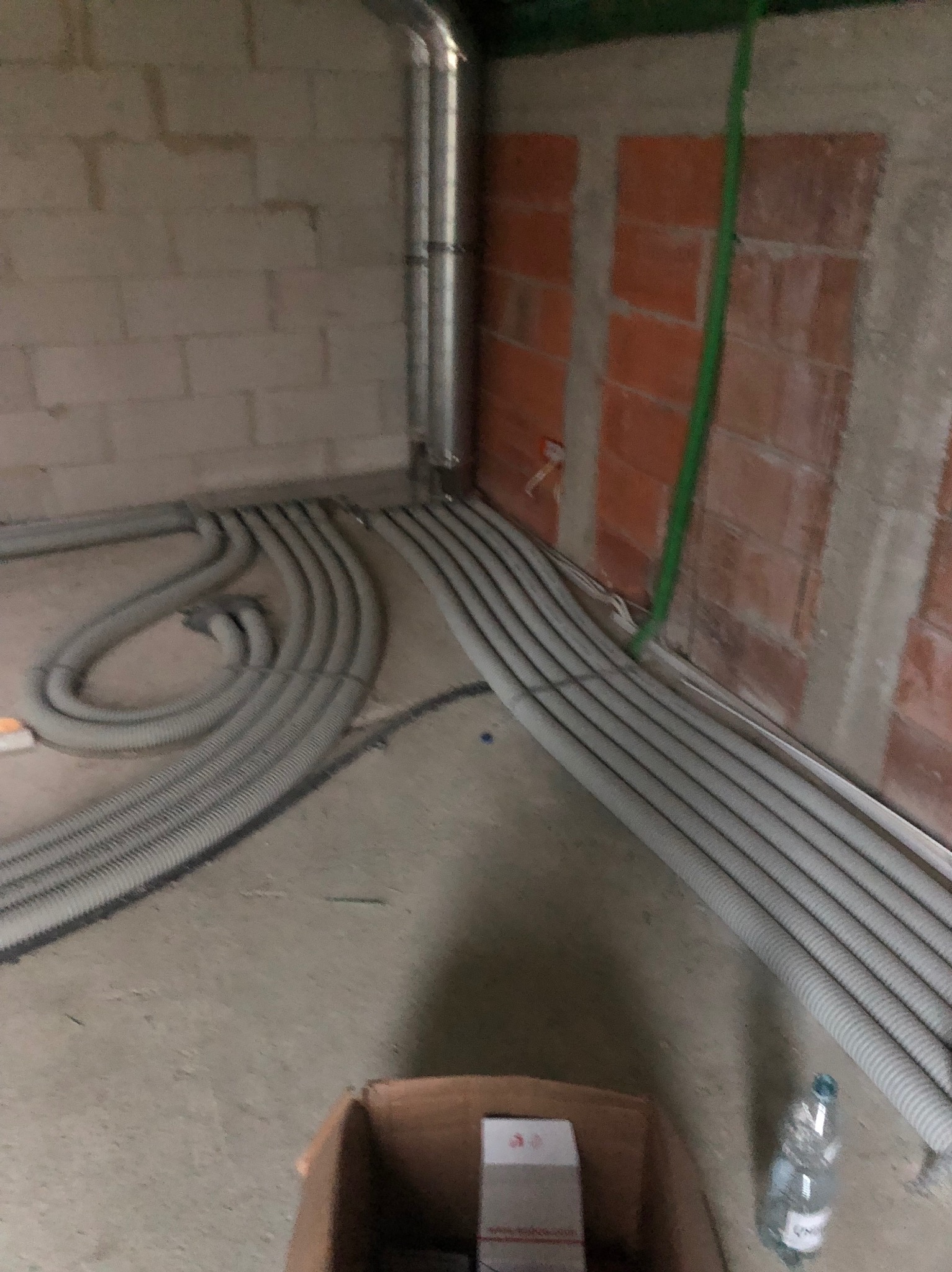 Szare, karbowane rury wentylacyjne ułożone na podłodze w surowym wnętrzu budynku z ceglanymi ścianami. Widoczna butelka wody i kartonowe pudełko.