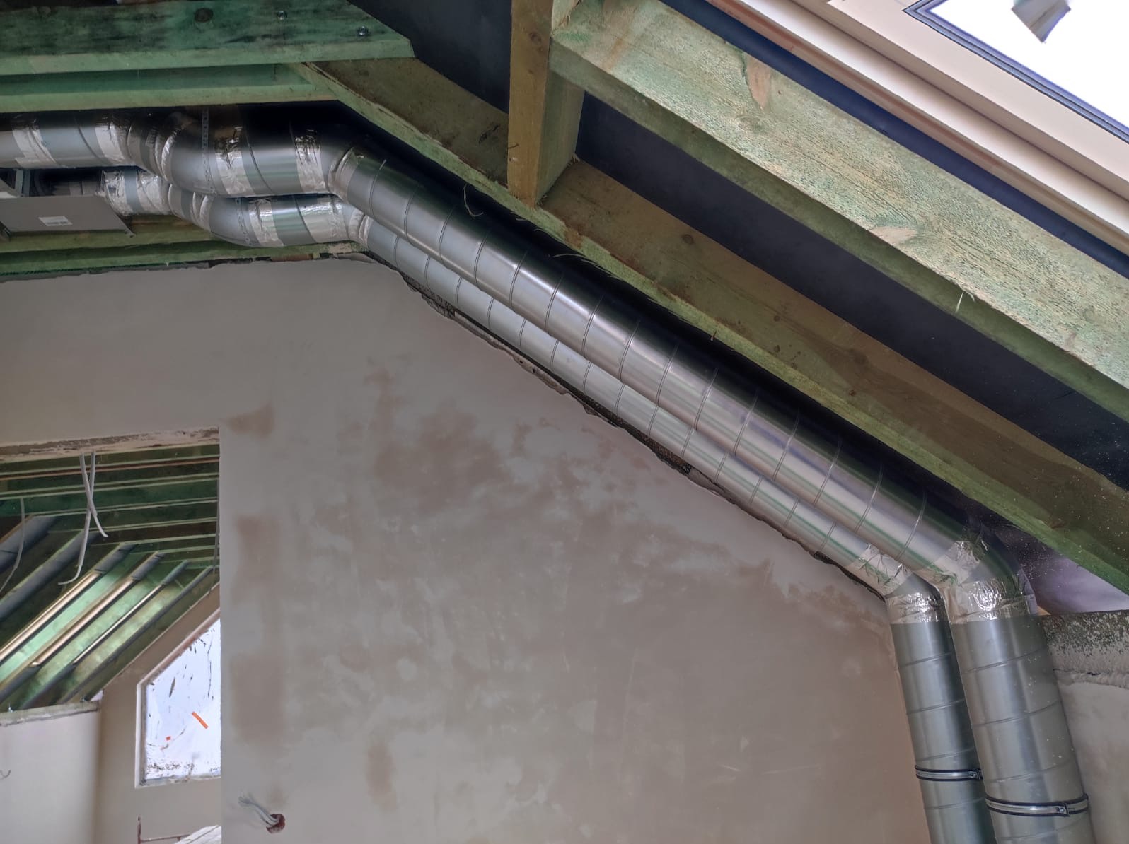 Dwa srebrne przewody wentylacyjne, owinięte taśmą, zamontowane pod drewnianą konstrukcją dachu na poddaszu w trakcie prac wykończeniowych.