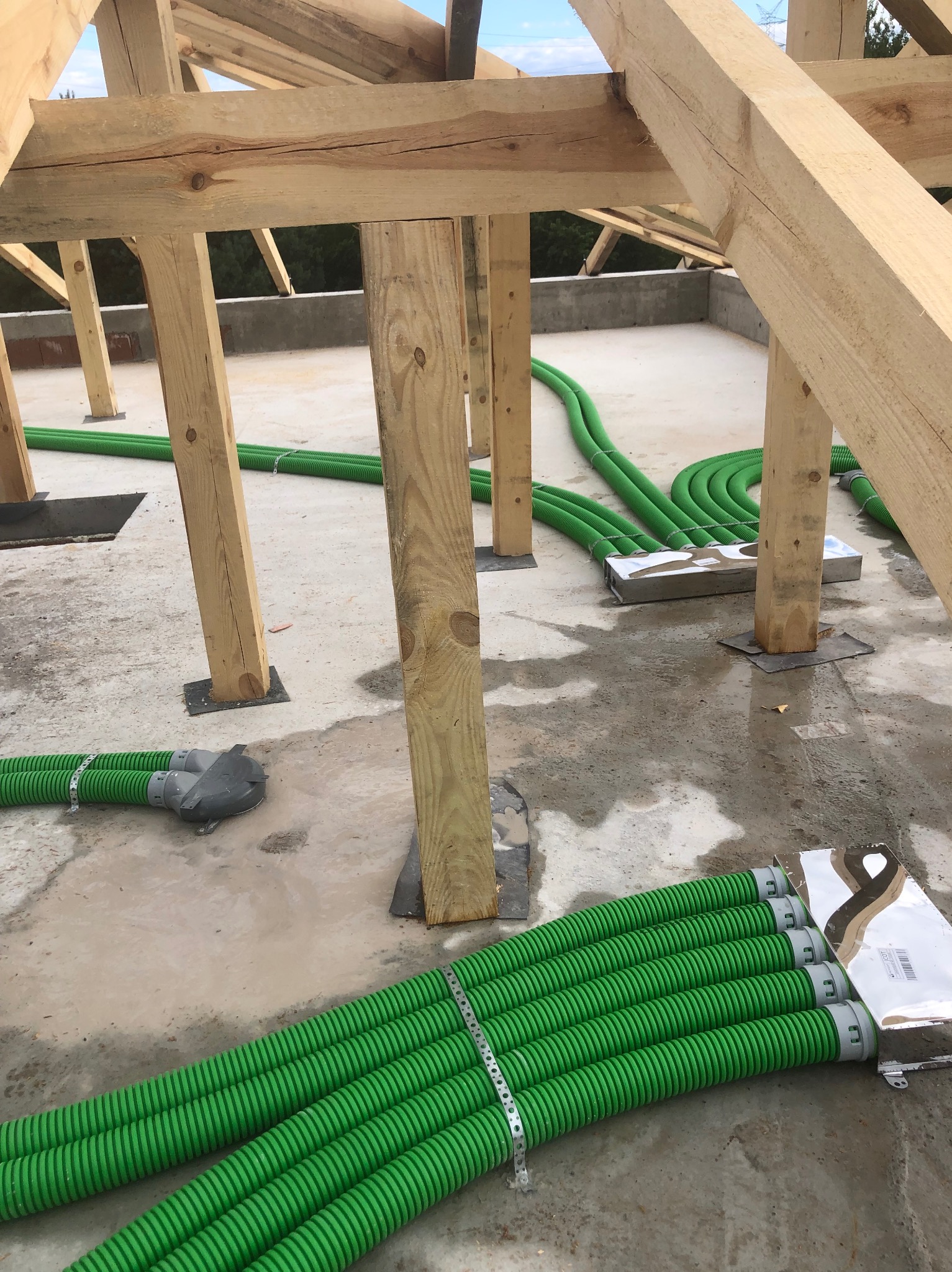 Surowe drewno konstrukcji dachu i zielone rury wentylacyjne na betonowej posadzce, przygotowane do montażu, z widocznymi elementami mocującymi i opakowaniem.