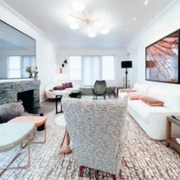 Elegancki salon z marmurowym kominkiem, nowoczesnym oświetleniem sufitowym i dywanem w geometryczne wzory, widziany zza fotela w szarej tkaninie.