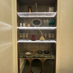 Fajny Elektryk Maciej Karaś - Wnęka ścienna z podświetlanymi półkami eksponująca szkło dekoracyjne i naczynia, z marmurowym blatem i szafkami z metalowymi wykończeniami.