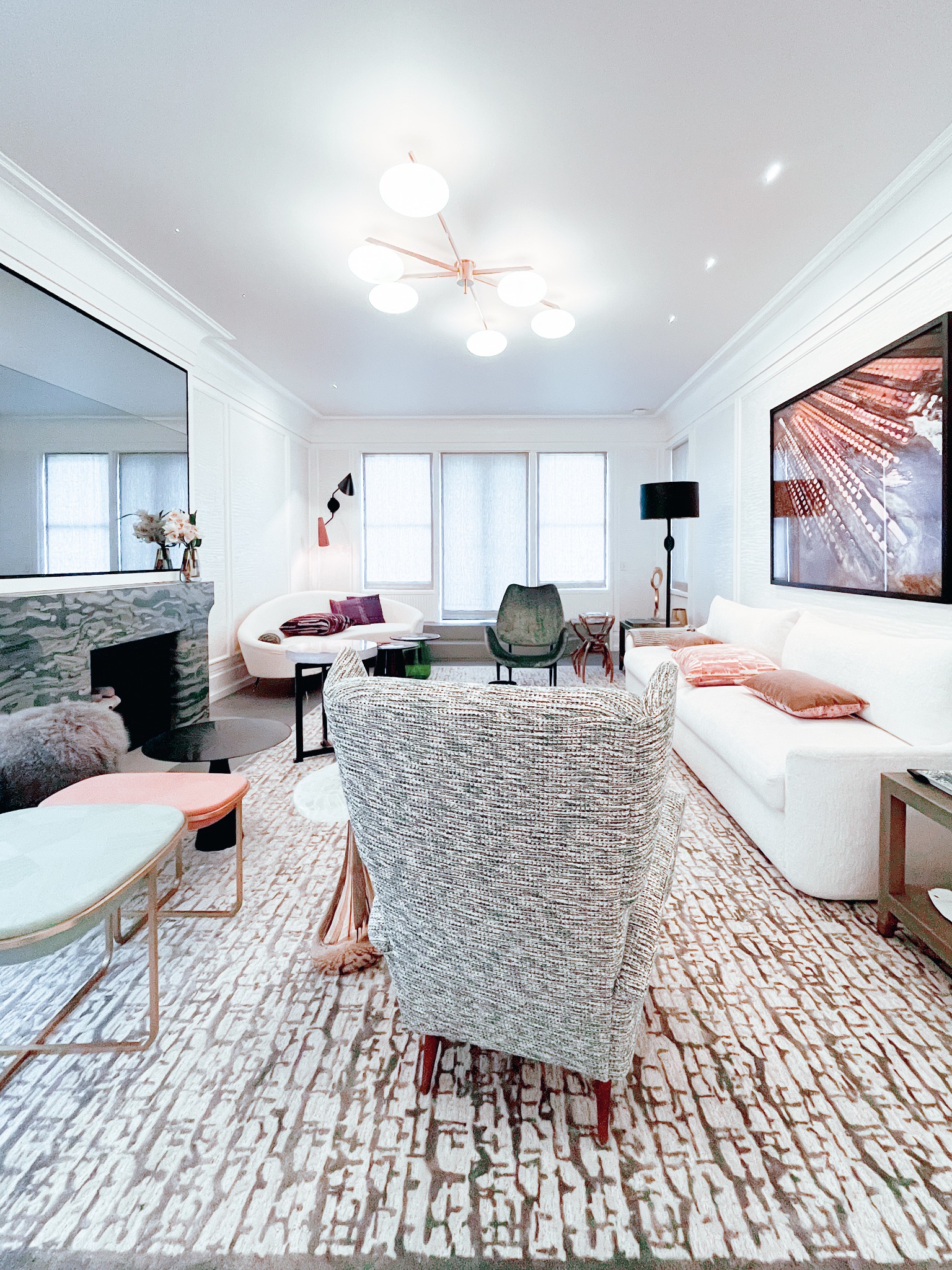 Elegancki salon z marmurowym kominkiem, nowoczesnym oświetleniem sufitowym i dywanem w geometryczne wzory, widziany zza fotela w szarej tkaninie.