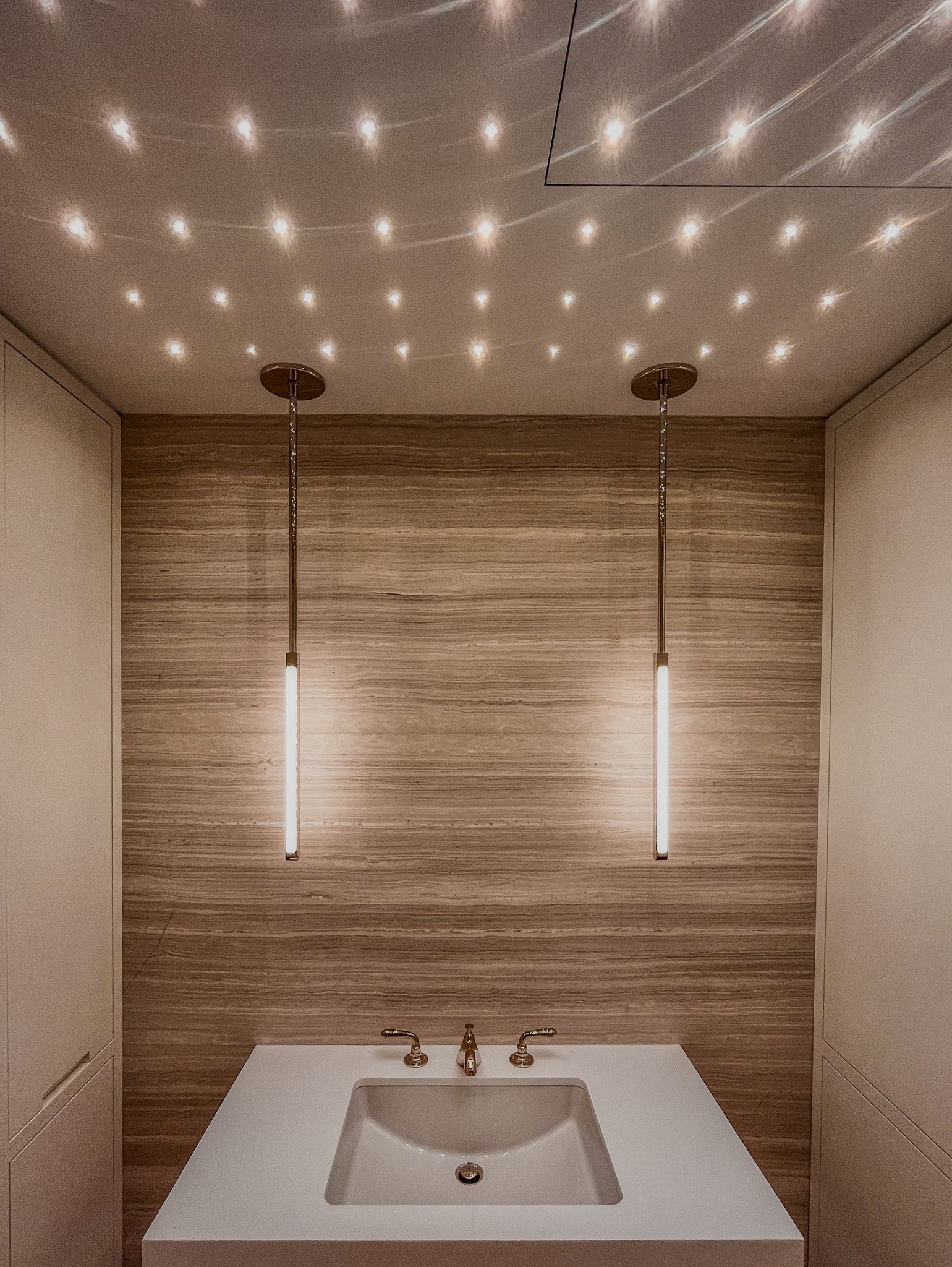 Nowoczesna łazienka z dwoma podłużnymi lampami wiszącymi nad umywalką, oświetlenie punktowe na suficie tworzy efekt gwiazd, ściana wykończona panelami imitującymi drewno.