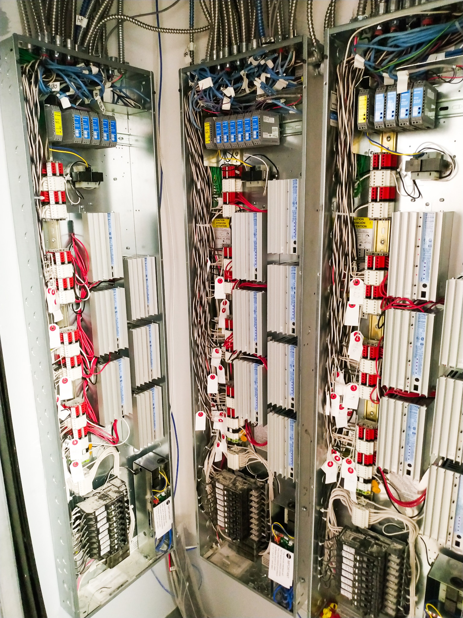 Trzy metalowe szafy elektryczne z widocznymi przewodami, bezpiecznikami, modułami sterującymi i innymi elementami, tworzące rozbudowany system zasilania i dystrybucji energii.