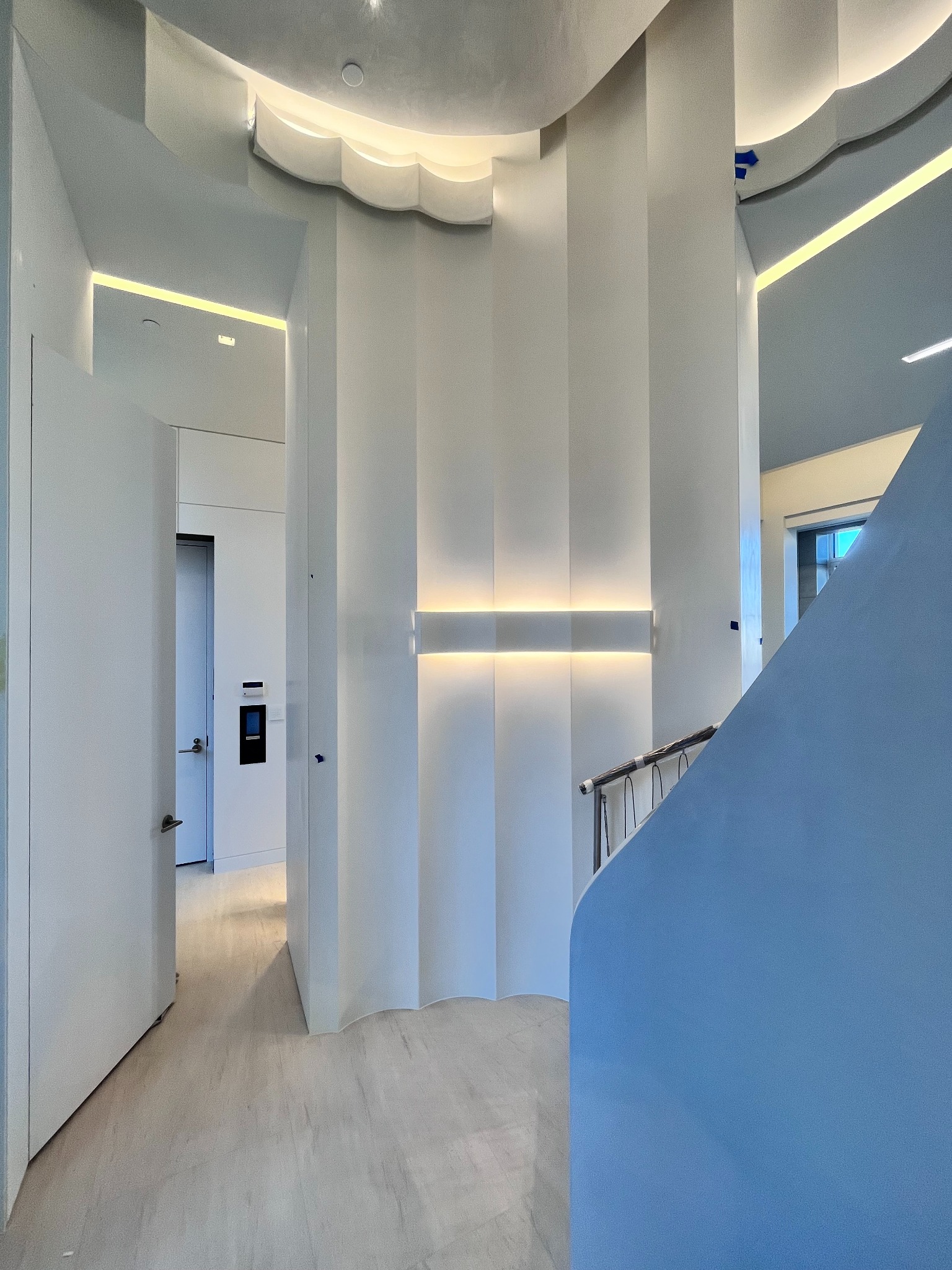 Nowoczesne wnętrze domu z białymi ścianami, oświetleniem LED w suficie i ścianach, otwartymi drzwiami do pomieszczenia z panelem sterowania, oraz fragmentem niebieskiej ściany schodów.