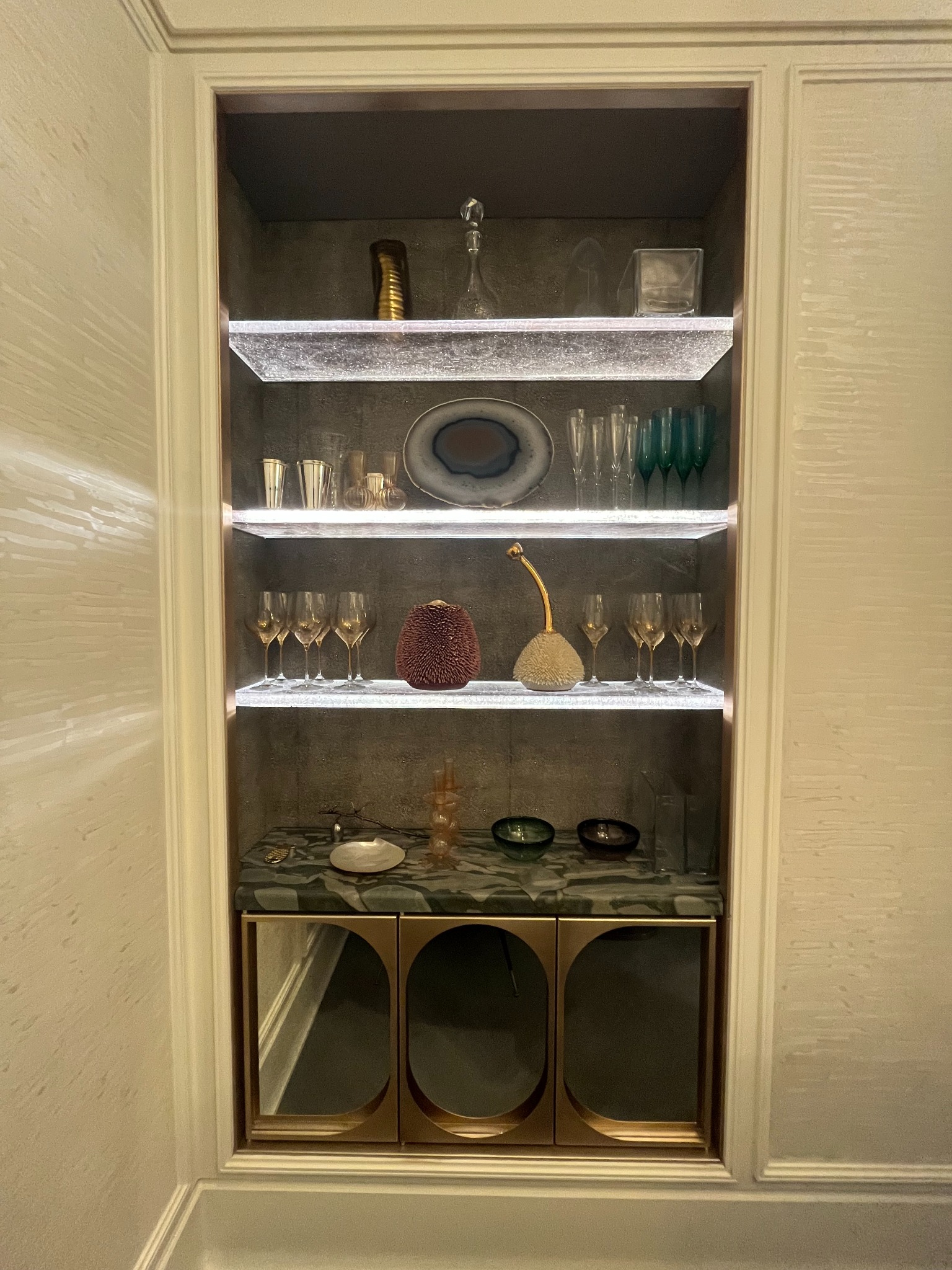 Wnęka ścienna z podświetlanymi półkami eksponująca szkło dekoracyjne i naczynia, z marmurowym blatem i szafkami z metalowymi wykończeniami.